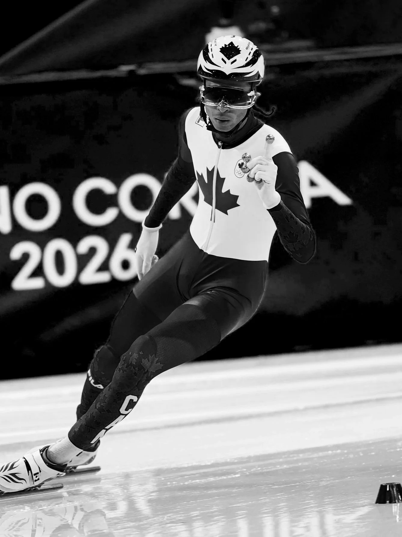 A skier wearing a Canadian team uniform, helmet, and goggles is racing on an ice track during the 2026 Olympics.