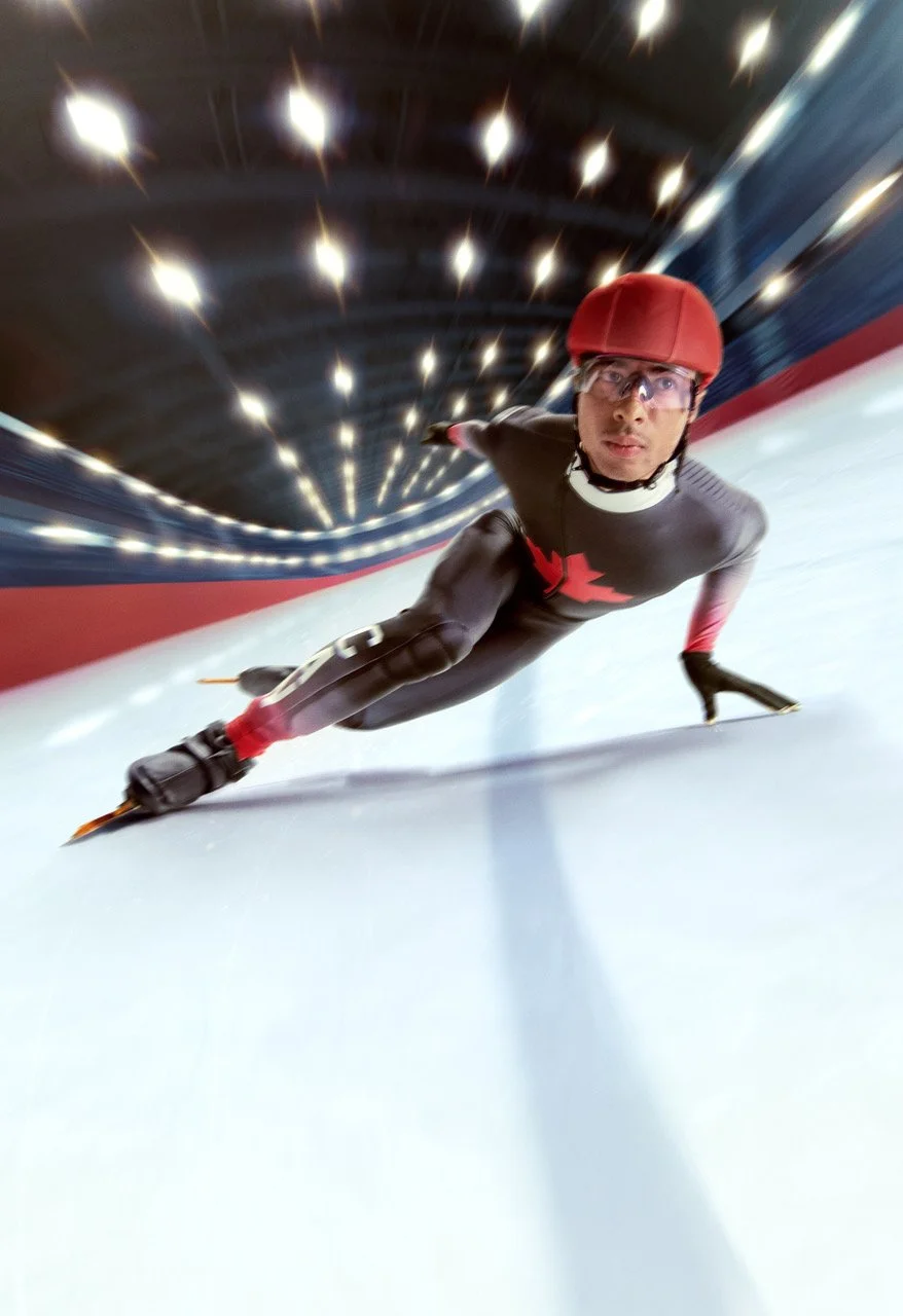 A person speed skating on an indoor ice rink, wearing a red helmet, glasses, and a black racing suit with red accents, captured mid-turn.