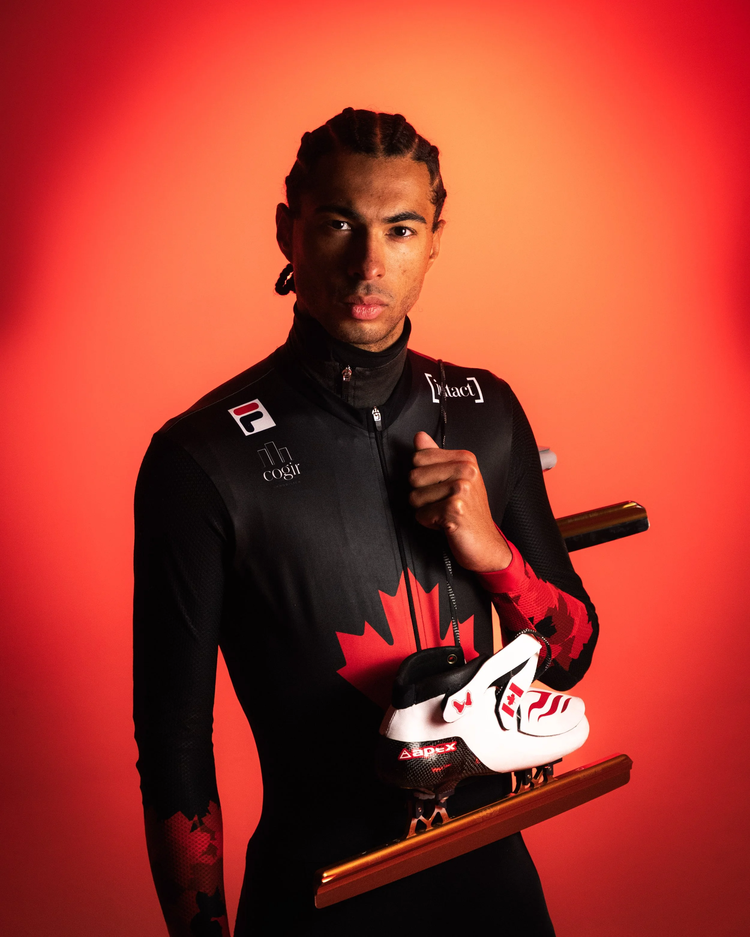 Portrait of a young man with braided hair, wearing a black and red sports outfit with sponsor logos, holding a trophy or award, against a red gradient background.