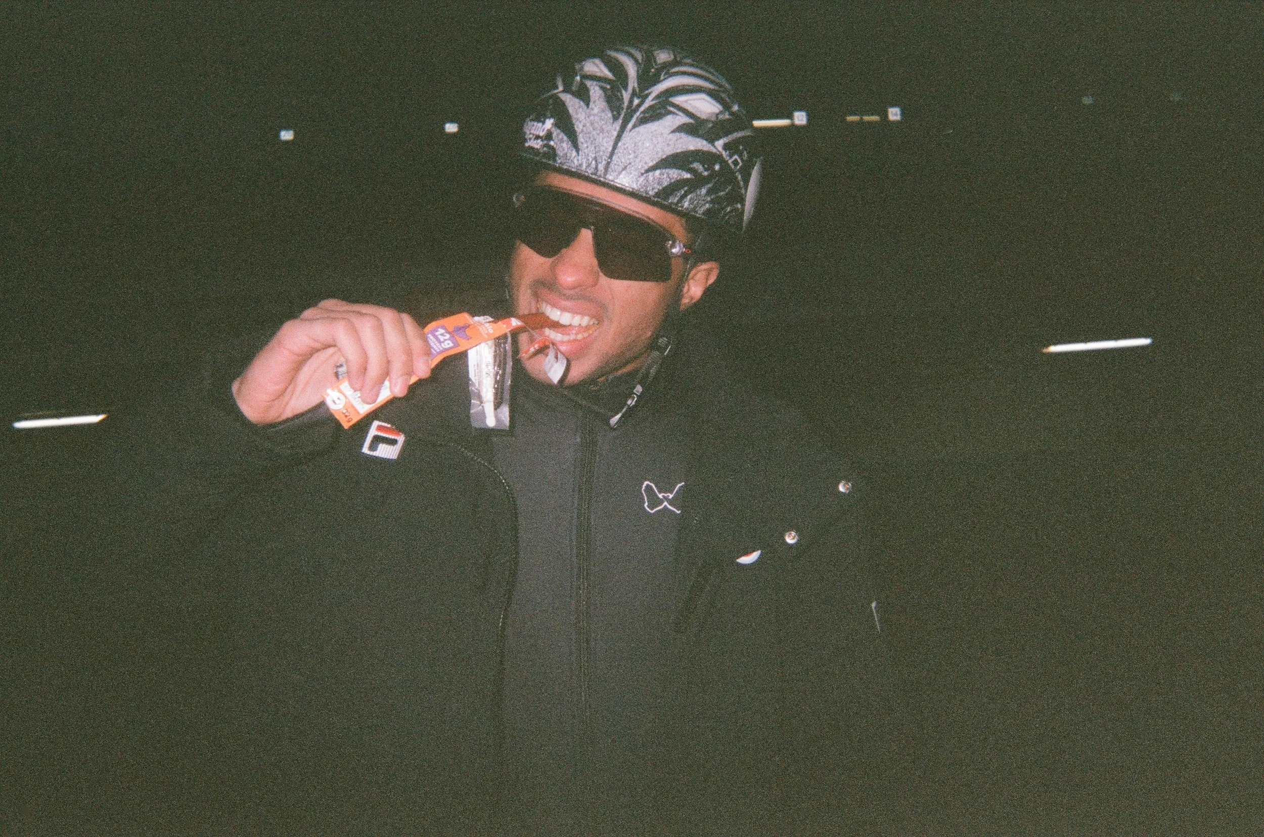 A person wearing a helmet and sunglasses is biting into a snack bar, smiling, in a dark environment.