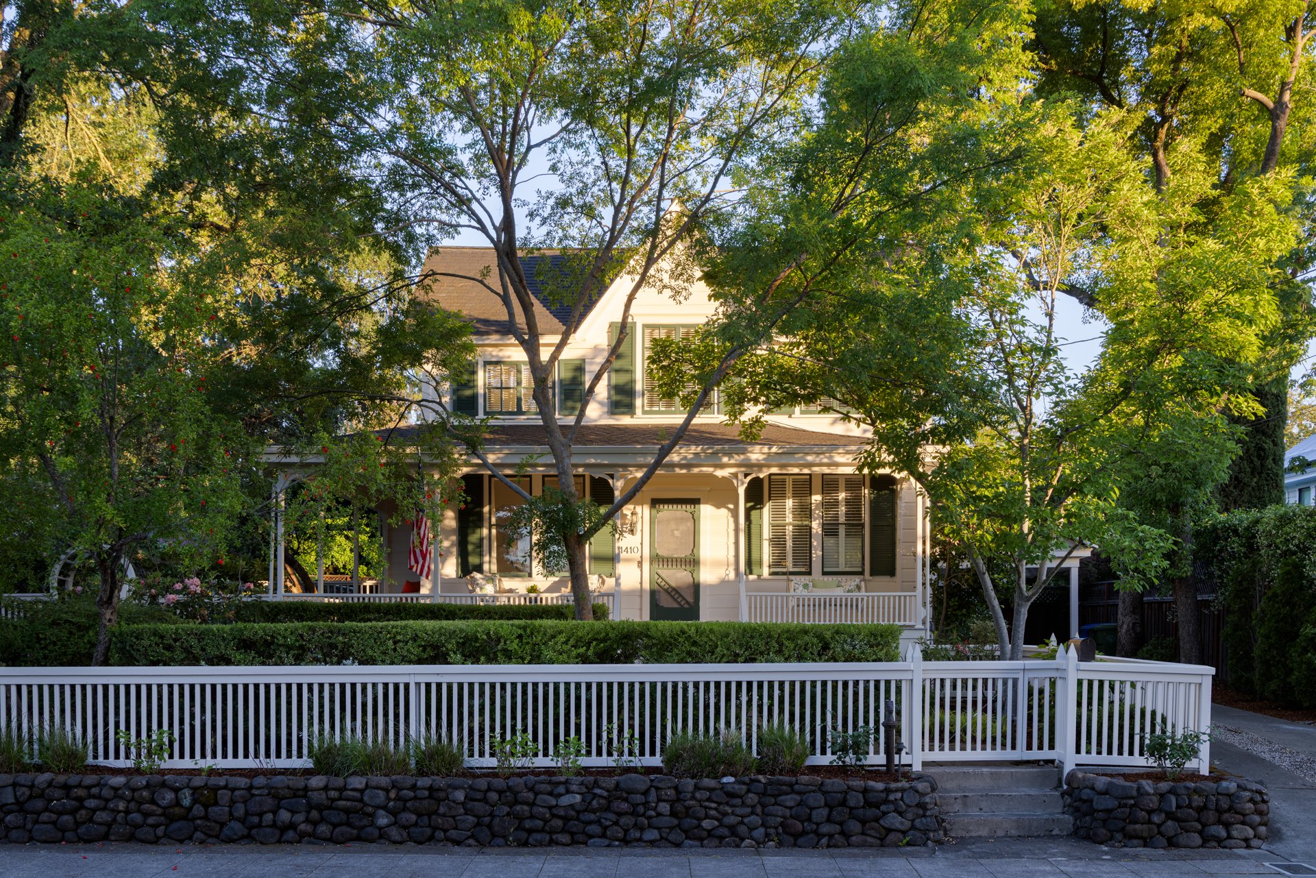 A white two-story house surrounded by trees with green leaves, a white picket fence, and a stone-lined garden bed in front.