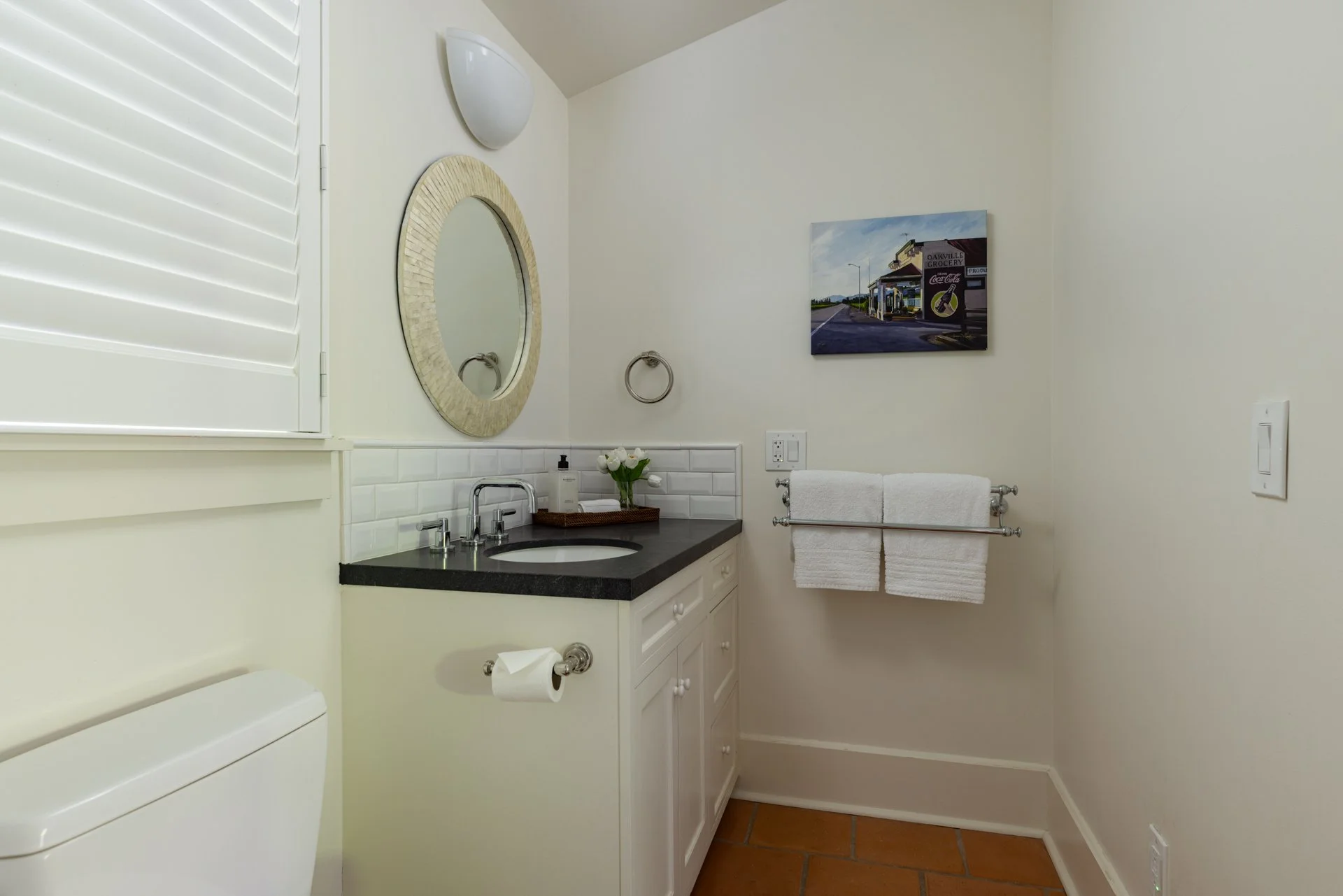 A bathroom with a white vanity, black countertop, oval mirror, wall-mounted light, towel ring with a white towel, and a framed picture of a gas station on the beige wall. There is a white toilet paper roll on a holder below the vanity and a white tow