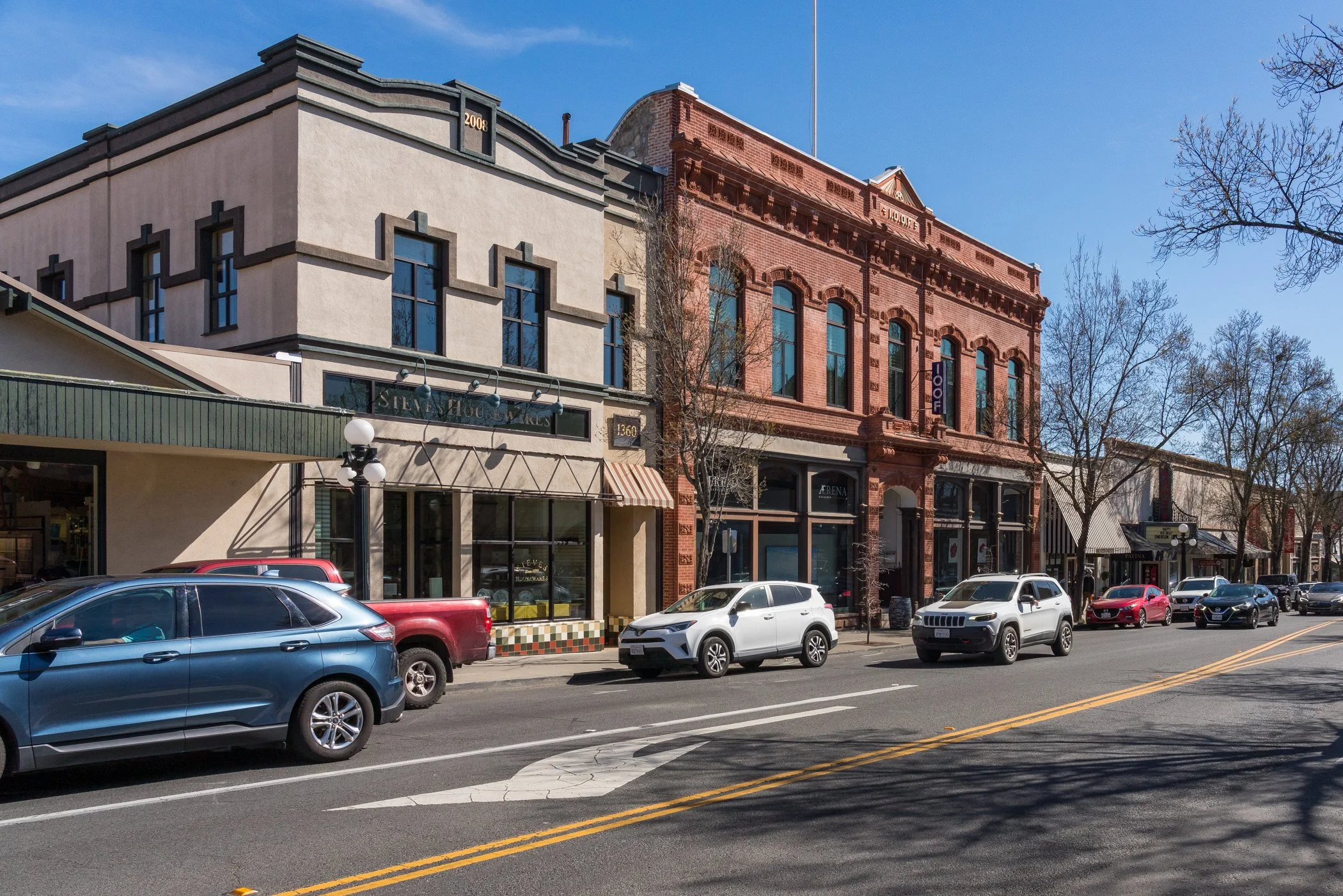 Street view of storefronts with parked cars along the curb, two buildings with different architectural styles, and leafless trees in front.