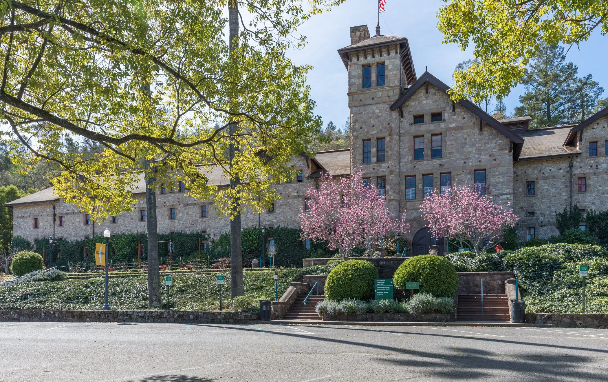 A large stone building with a tower-like structure surrounded by trees, pink flowering trees, and landscaped bushes in the foreground.