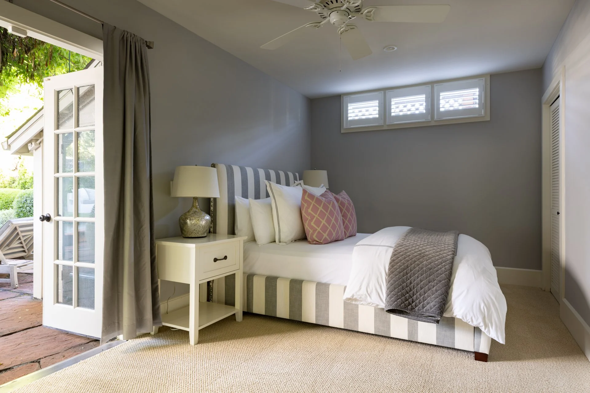 A bedroom with a bed, pillows, a bedside table with a lamp, a ceiling fan, windows with shutters, an open door with curtains revealing an outdoor patio with greenery.