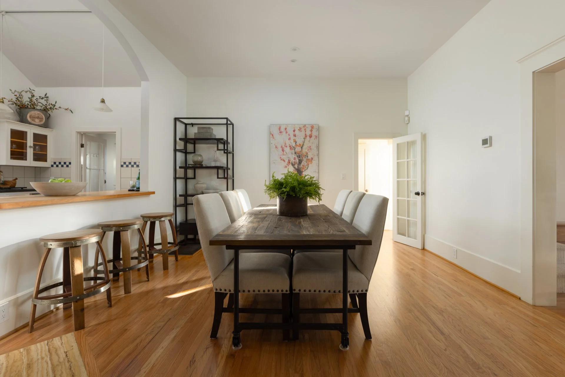Dining area with a wooden table, cushioned chairs, a black metal shelving unit, and artwork on the wall. An open door leads outside, and the kitchen with bar stools is partially visible.