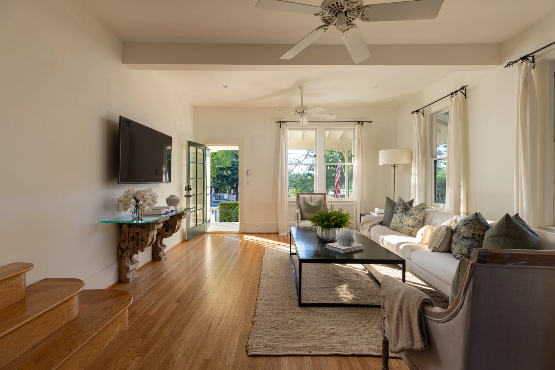 Living room with white walls, hardwood floor, reclining couch with decorative pillows, coffee table, floor lamp, wall-mounted TV, front door with glass panels, and windows with white curtains.