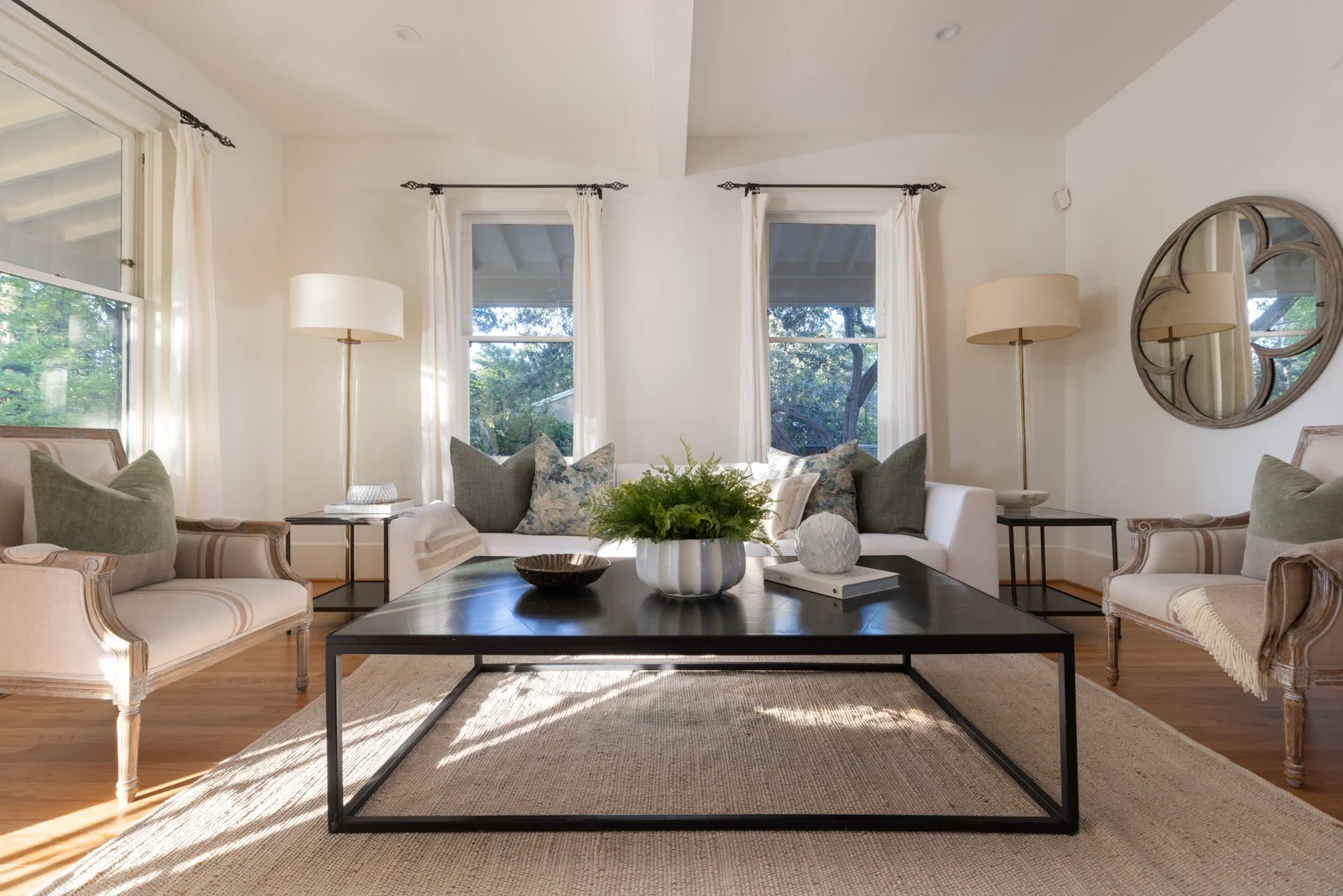 Living room with three large windows, white curtains, two white sofas with pillows, a dark wooden coffee table with decorative items, and tall floor lamps, a round mirror on the wall, and wooded flooring.