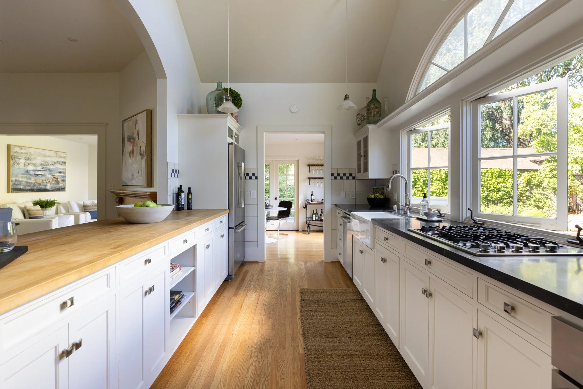 Kitchen with large arched window, white cabinets, black countertops, wooden island, stainless steel refrigerator, hardwood floor, and decorative bottles on high shelves.