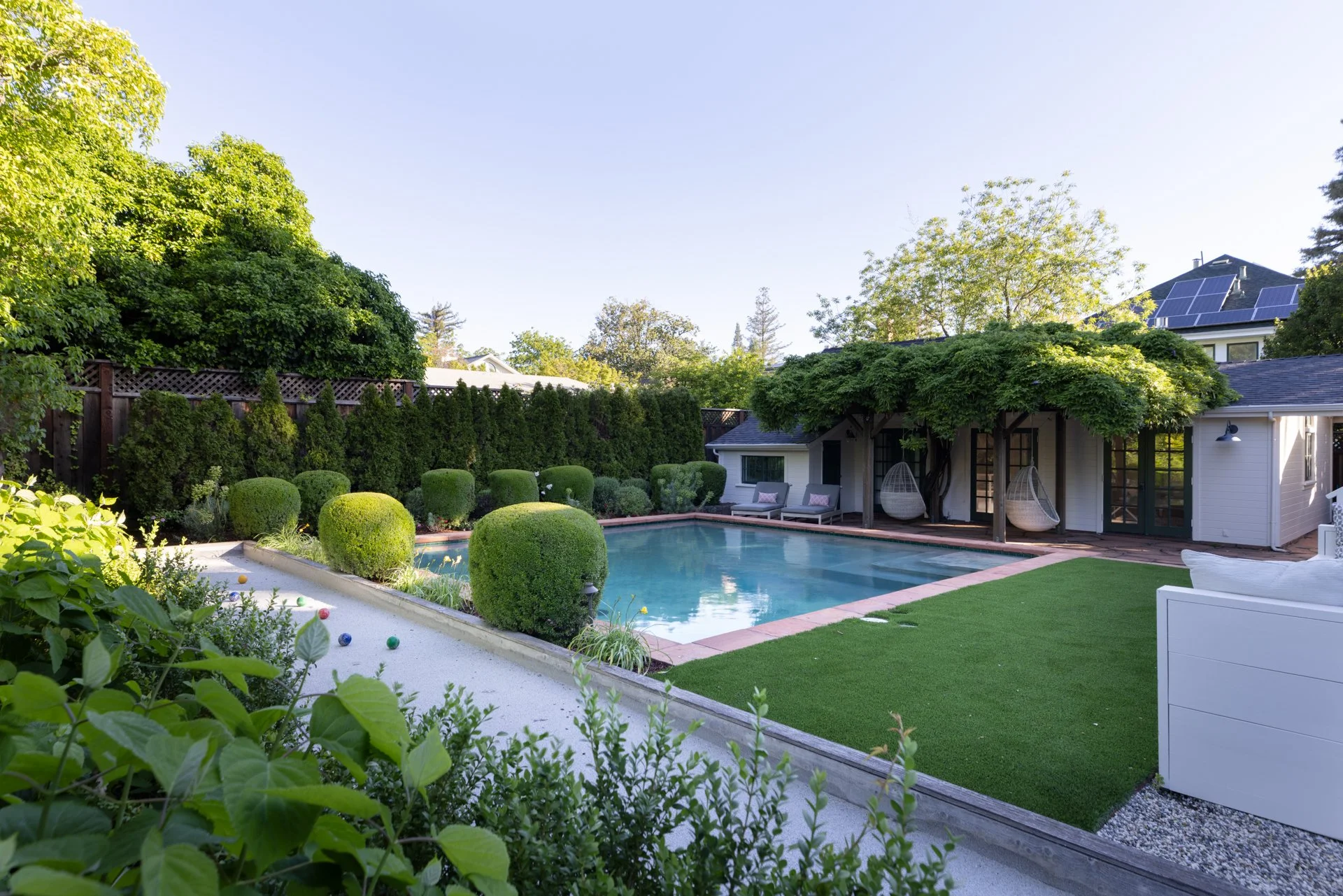 A backyard with a small swimming pool, surrounded by manicured bushes, trees, and a grassy area. There are lounge chairs by the house, which has solar panels on the roof, and a patio area with hanging outdoor chairs.