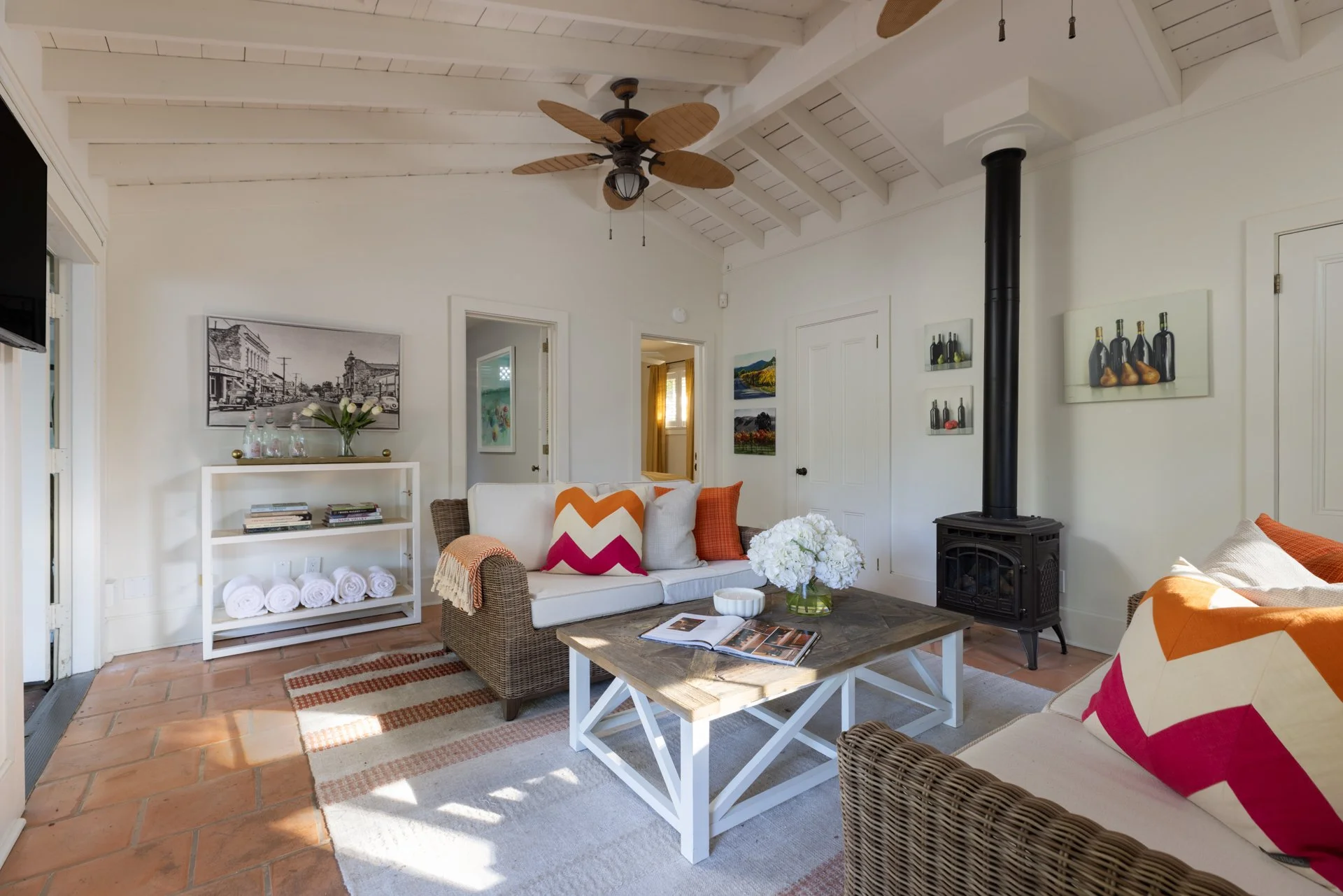 A cozy living room with white walls and a white wooden ceiling. There are colorful pillows on rattan-style sofas, a wooden coffee table with magazines and a vase of white flowers, and artwork on the walls. A black wood stove and a ceiling fan complet