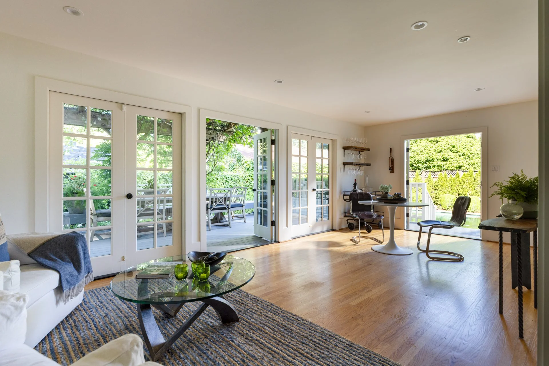 A bright living room with French doors opening to a green outdoor space, featuring a glass coffee table with green glasses, a white sofa with a blanket, and minimalistic decor.