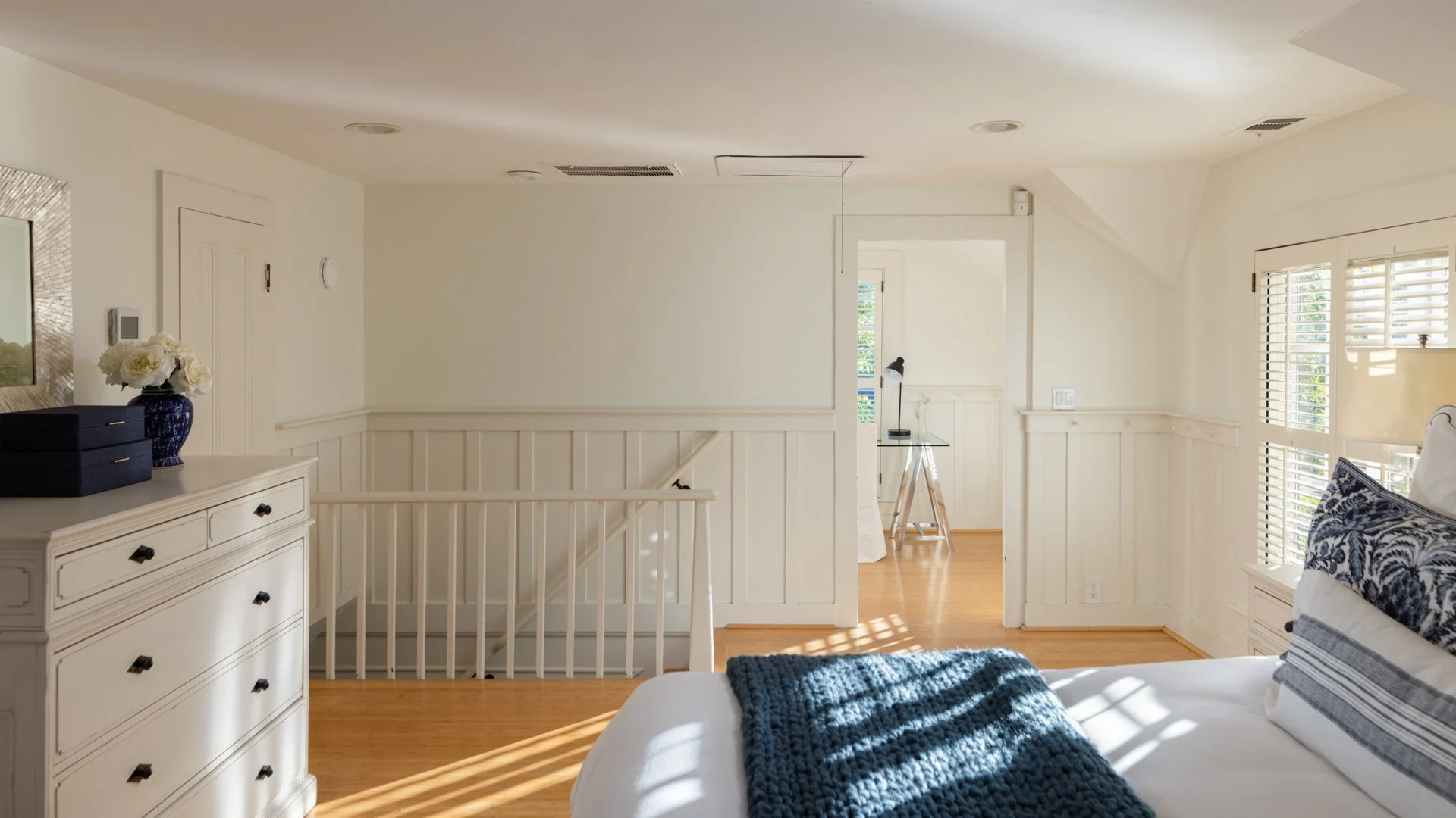 Bright and airy bedroom with white walls, large windows with shutters, and wooden flooring. A white dresser with black handles, decorated with a blue vase filled with white flowers and dark boxes, is on the left. To the right, a bed with white beddin
