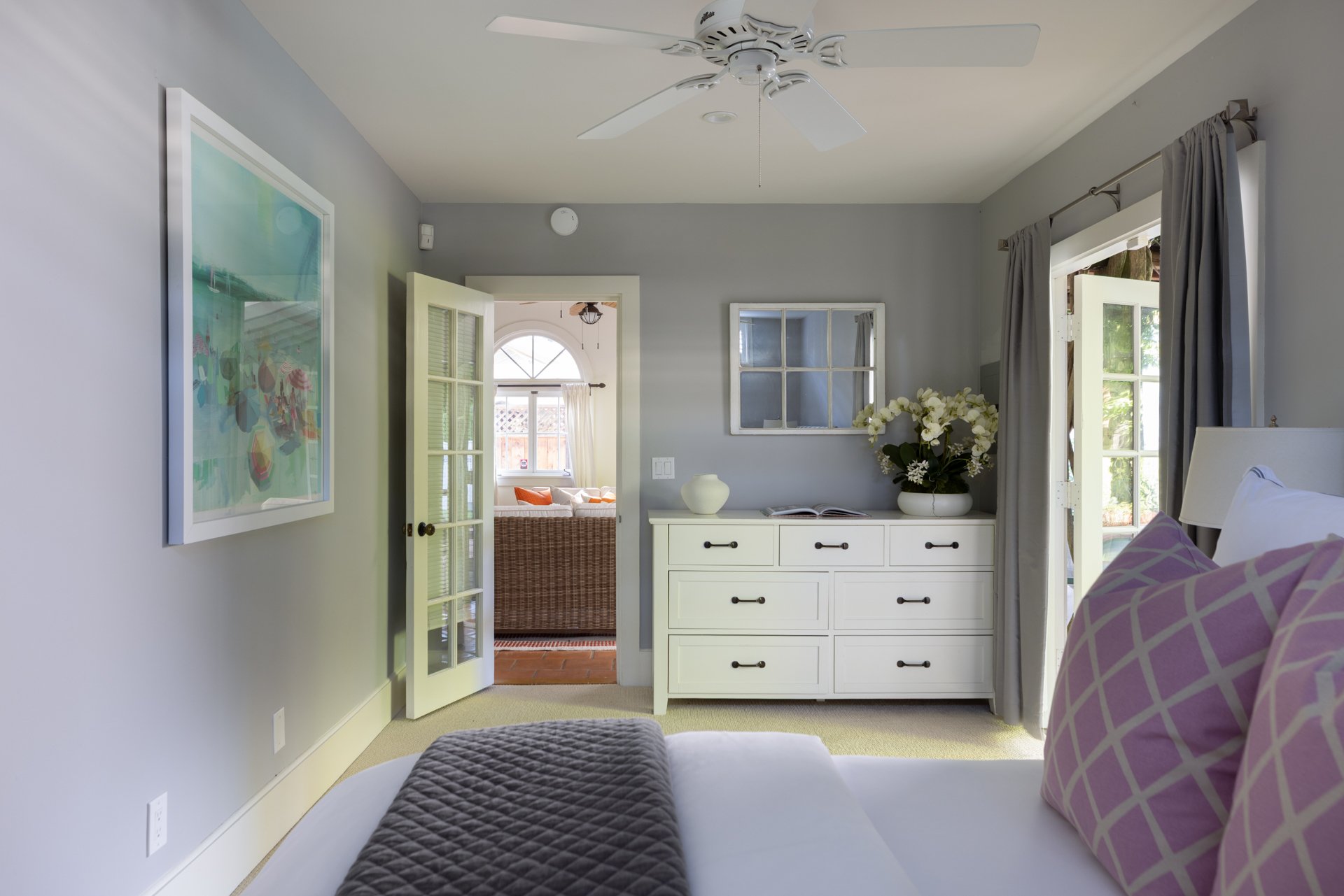 A bedroom showing part of a bed with purple pillows and a brown quilt, a white dresser with black handles, a vase with white flowers, door with glass panes leading to another room, a window with curtains, and a ceiling fan.