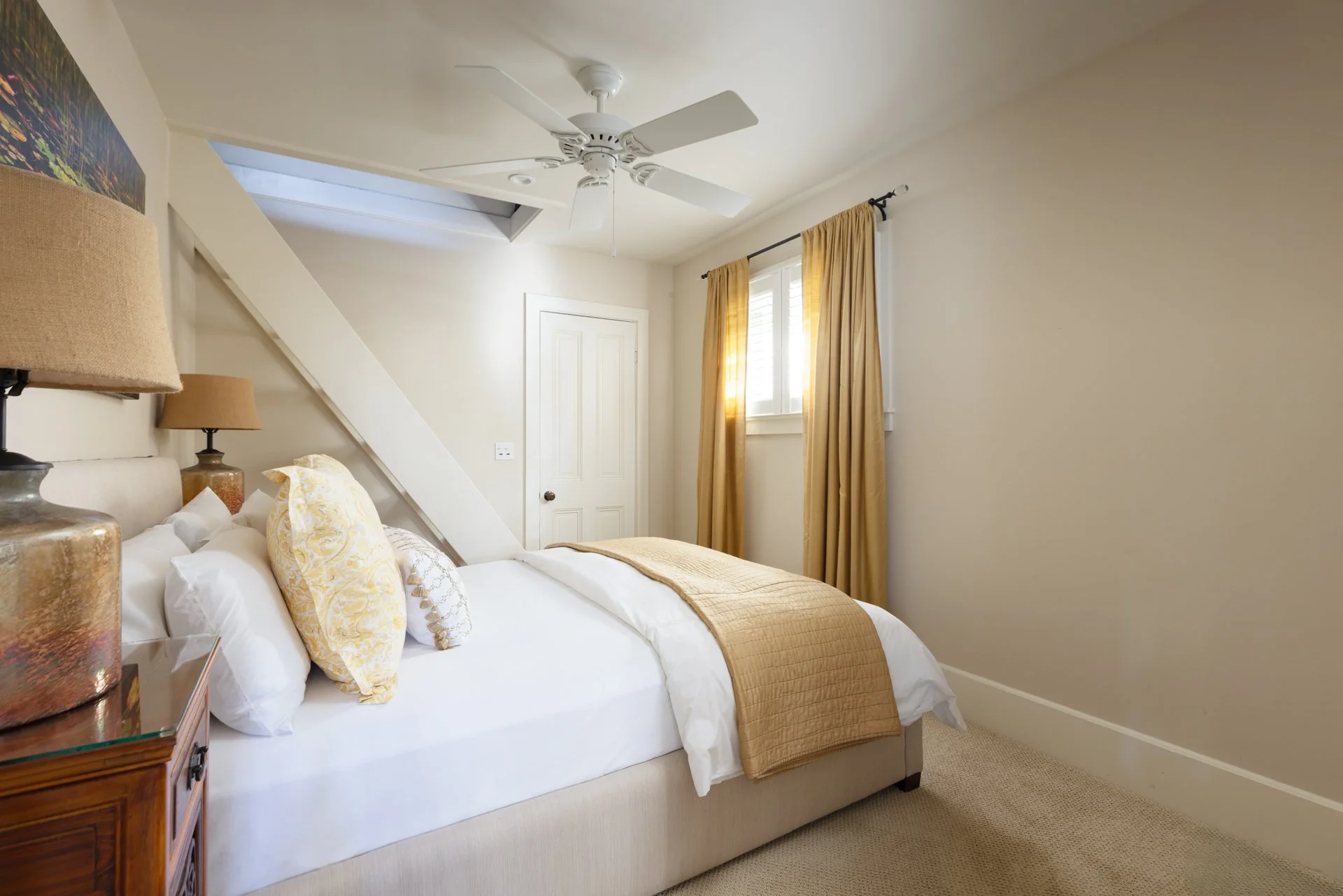 A cozy bedroom with a large bed covered with white and beige bedding, decorative pillows, a wooden nightstand with lamps, a white ceiling fan, a skylight, a window with yellow curtains, and a beige carpet.