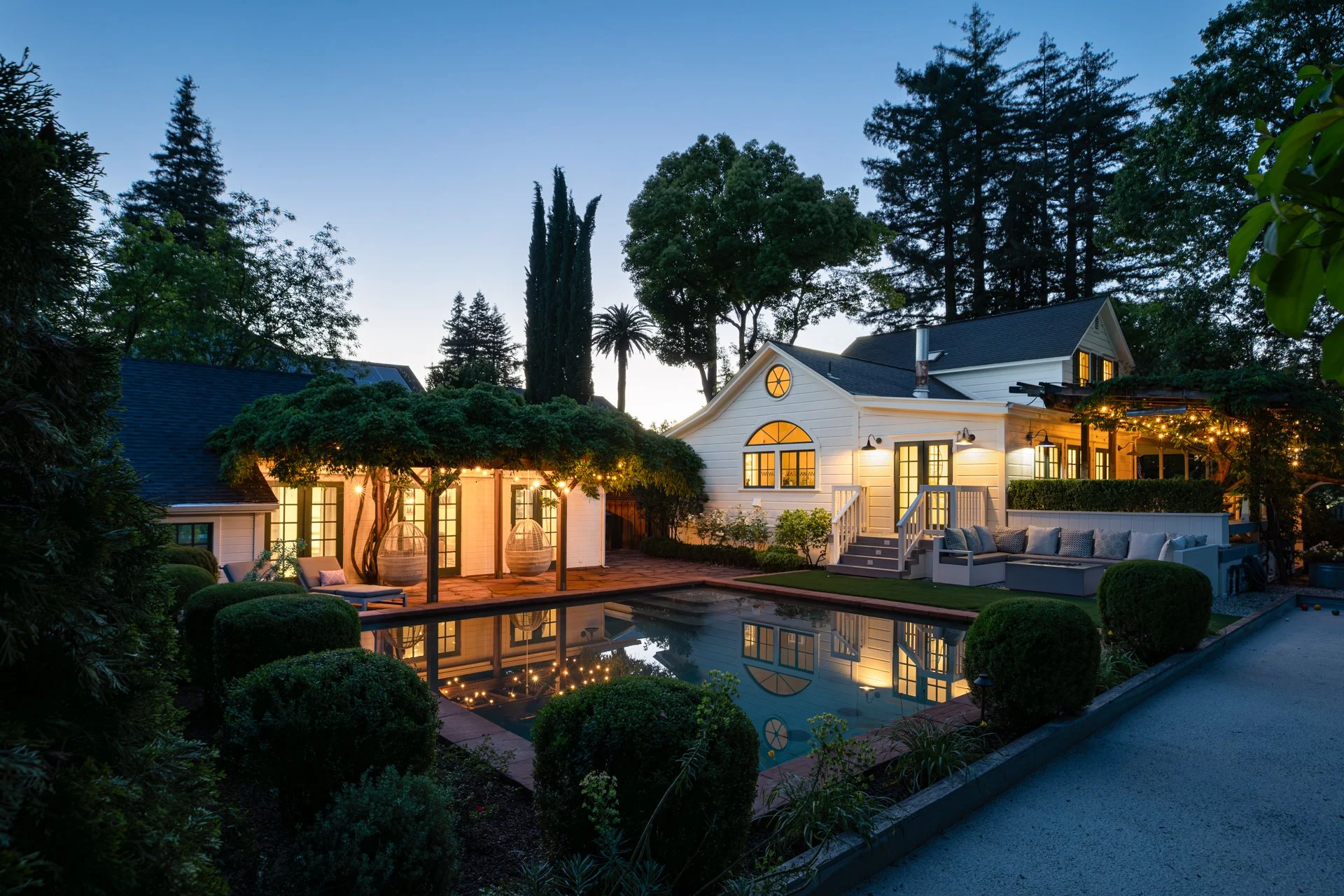 A backyard during dusk featuring a swimming pool reflecting the surrounding trees and house. The house has large windows and a patio with outdoor seating, illuminated by string lights and outdoor lamps. Tall trees and lush greenery surround the yard.