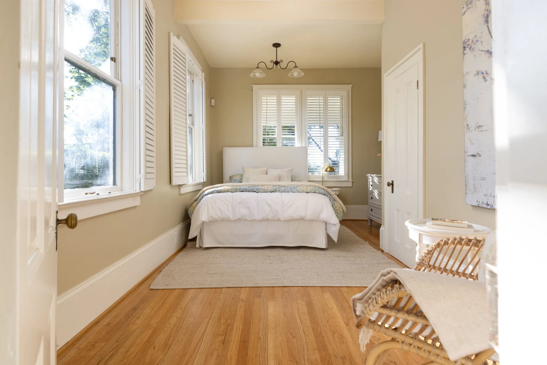 A cozy bedroom with a white bed, beige walls, multiple windows with white shutters, a beige rug, and woven chairs near the door, illuminated by natural light.