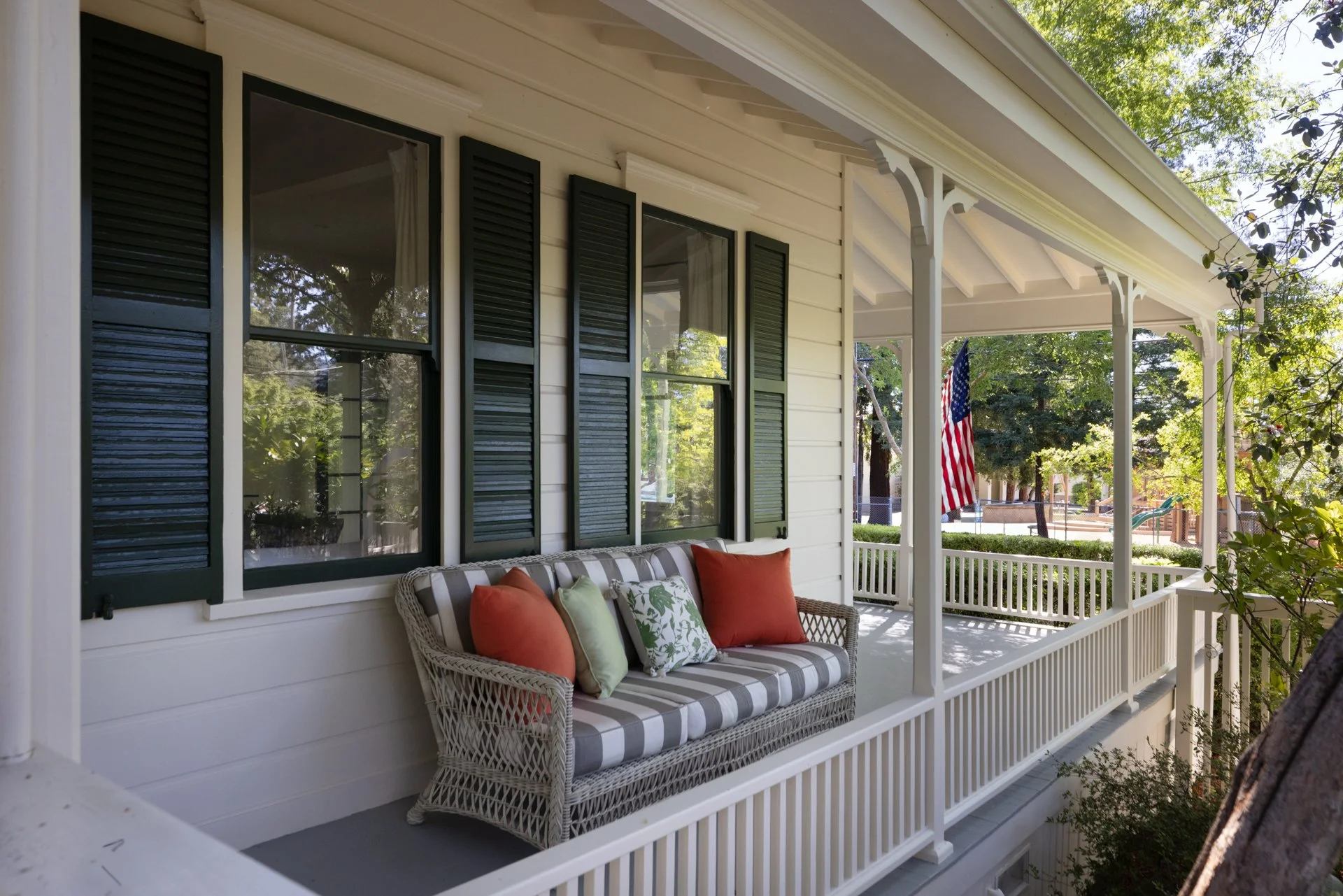 A front porch of a house with white siding and black window shutters, decorated with a wicker bench with colorful pillows, a hanging American flag, and surrounded by trees and greenery.