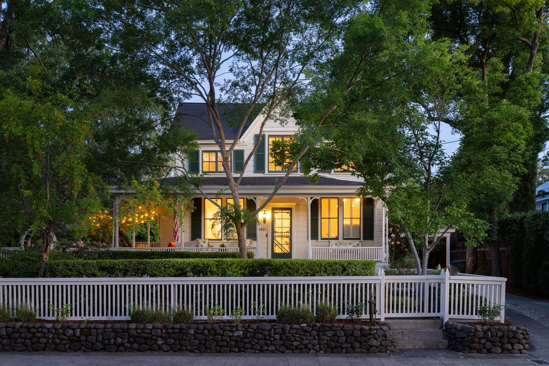 A two-story white house with black shutters and a front porch, illuminated from inside, surrounded by trees and greenery, with a white picket fence in front.