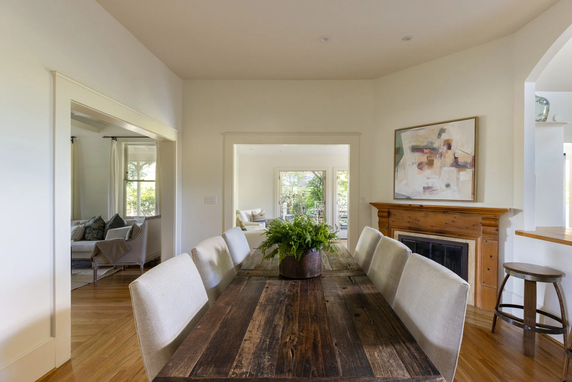 View of a dining room with beige upholstered chairs around a rustic wooden table, a potted plant centerpiece, a fireplace with wooden mantle, an abstract painting on the wall, and a living room visible through doorways with chairs and windows with cu