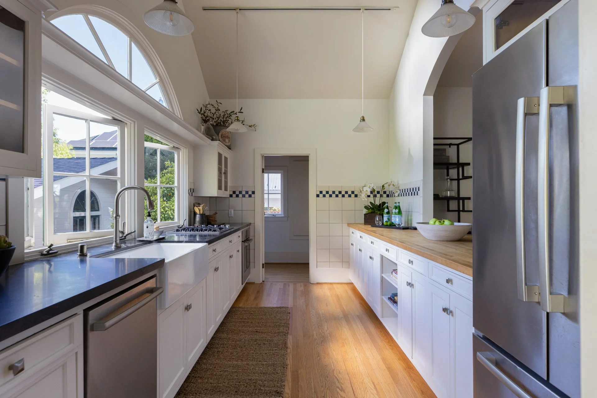 Bright kitchen with large arched window, white cabinets, wooden countertops, stainless steel refrigerator, and decor including plants and green apples.
