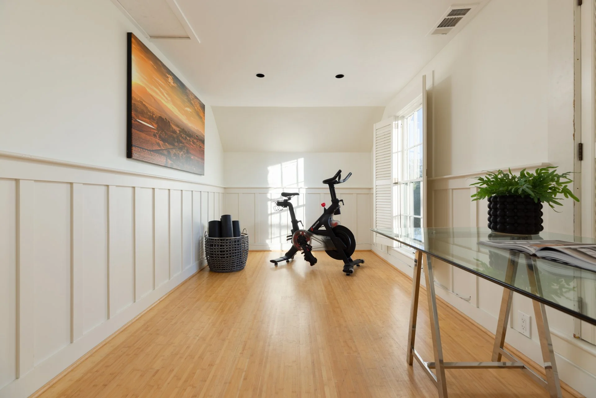 A bright living room with white walls, wooden floors, large windows with shutters, a black exercise bike, a basket with yoga mats, a glass table with a potted plant and magazines, and a colorful landscape painting on the wall.