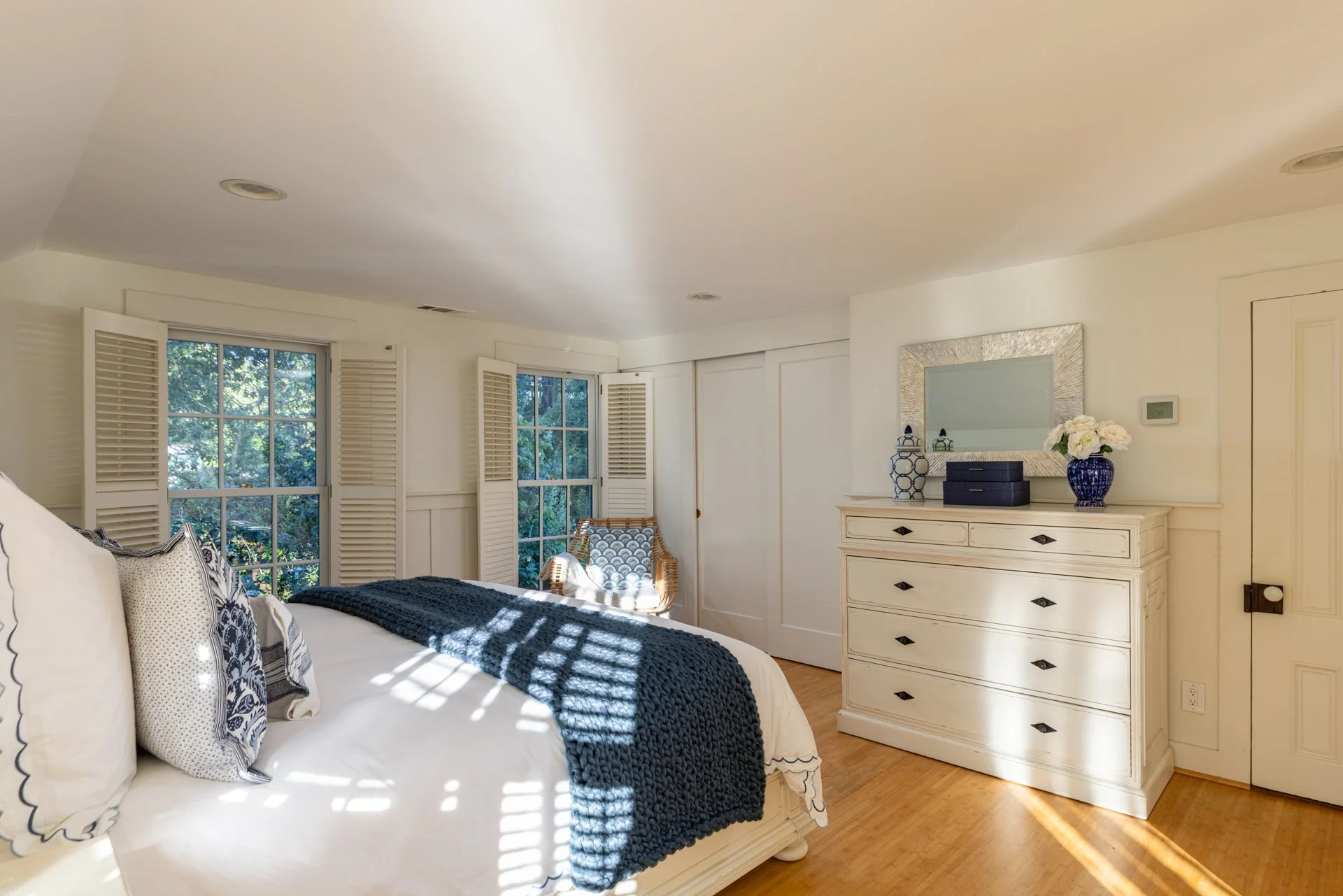 A cozy bedroom featuring white walls and a large window with plantation shutters, decorated with patterned pillows on a bed, a wicker chair, and a white dresser topped with vases and a mirror, with sunlight streaming through.