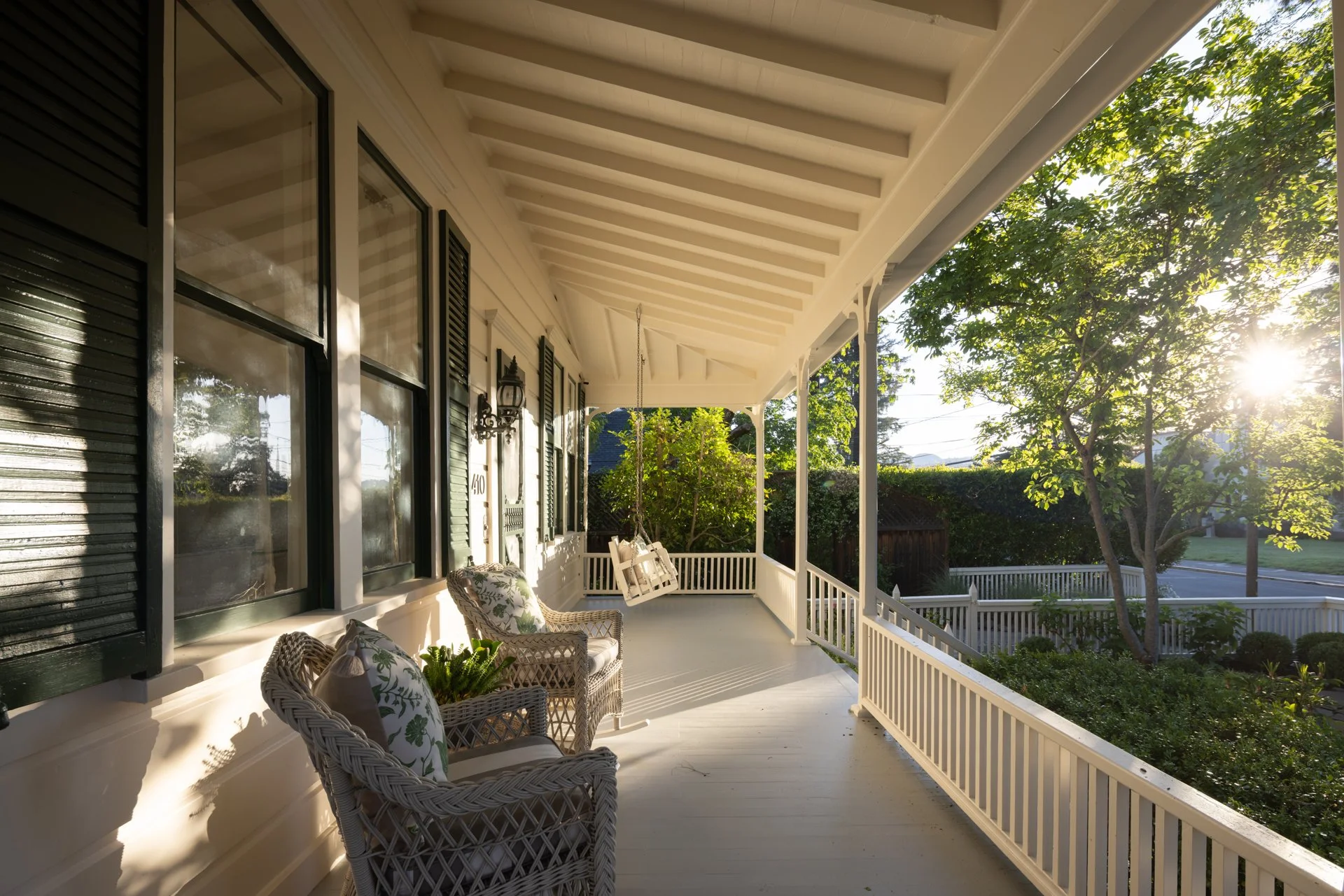 Front porch with wicker chairs and floral cushions, white railing, green trees, and sunlight shining through the leaves.
