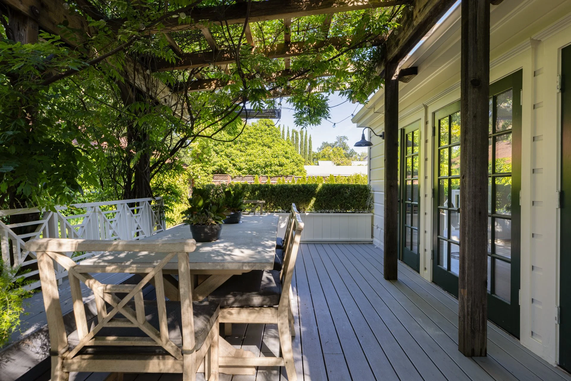 A wooden outdoor patio with a table and chairs, surrounded by green trees and plants, with a house wall on the right side featuring windows and a door, and a hedge in the background.