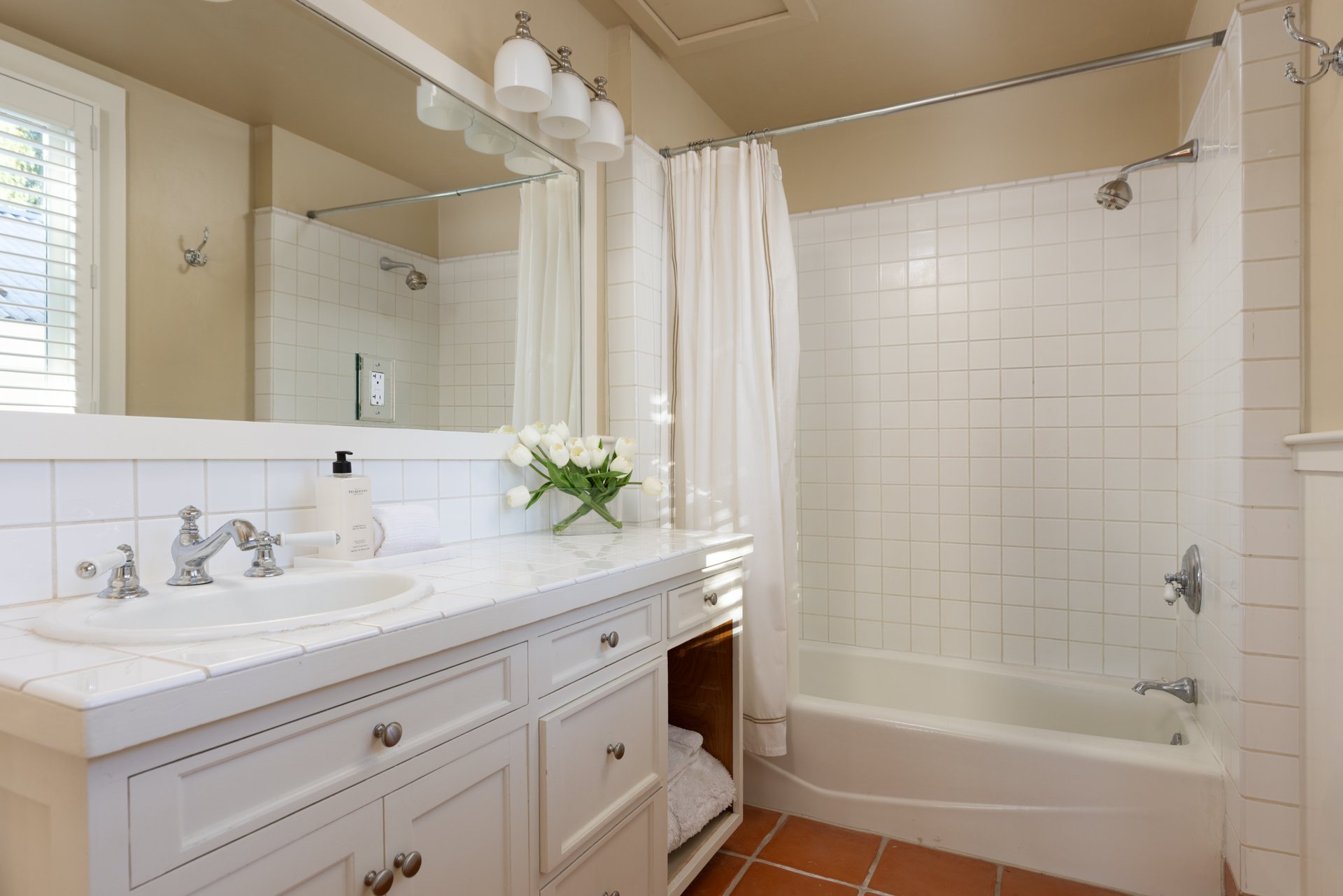 Bathroom with white tiled shower area and white vanity countertop, decorated with a vase of white flowers, and a large mirror above the sink. There is a window with blinds on the left side.