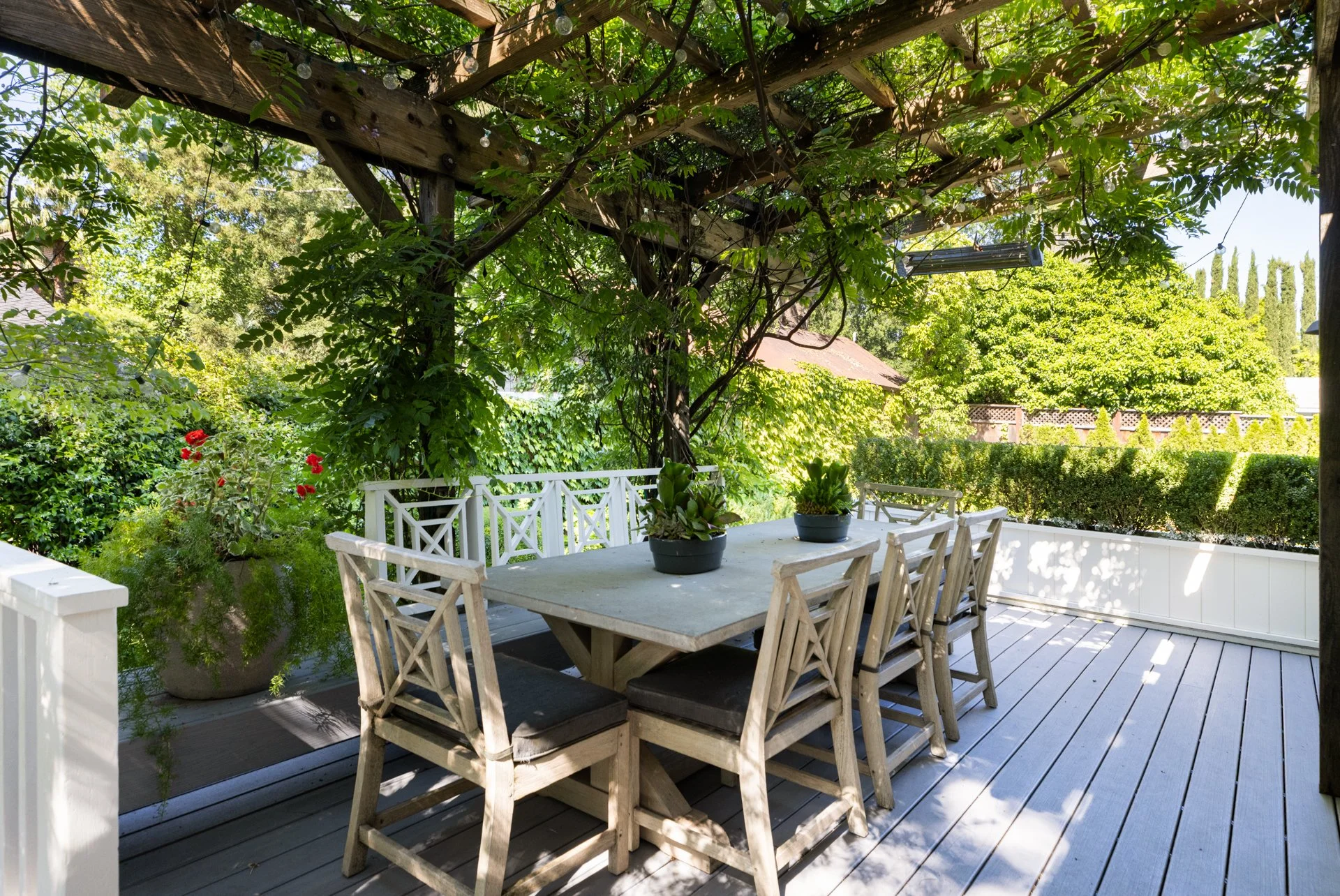 An outdoor patio decorated with lush green trees and plants, including red flowers in pots. There is a white table with matching chairs under a wooden pergola covered with vines and string lights, overlooking a garden with hedges and tall trees.