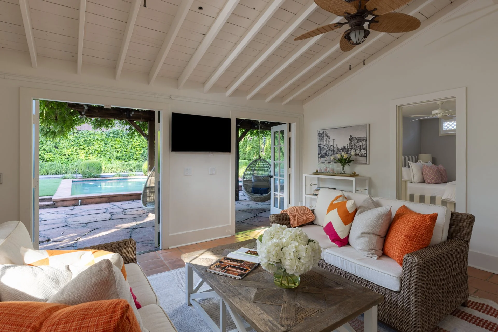 A cozy living room with a view of a backyard pool area through open French doors. The room has a white ceiling with exposed beams and a ceiling fan, beige walls, and modern decor, including a glass-top coffee table with a vase of white flowers, a whi