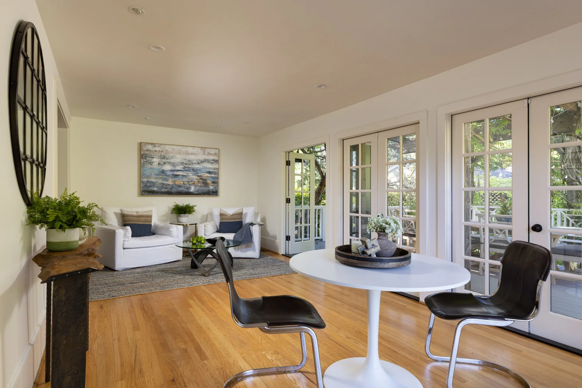 Living room with white walls, hardwood floors, and large windows opening to a balcony with greenery. Features include white sofas, a glass coffee table, wall art with abstract landscape, potted plants, and a round dining table with black chairs. The 
