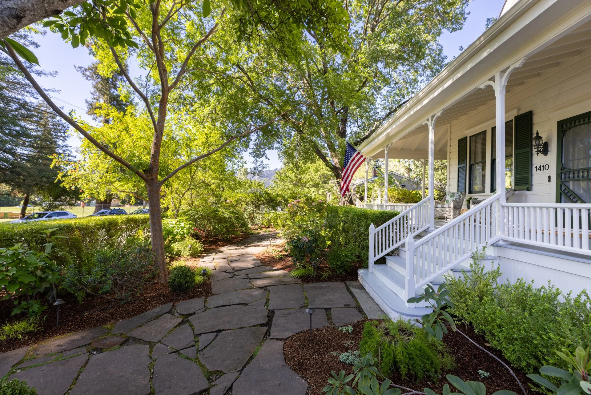 A front porch of a house with a white railing, steps, and a porch swing, surrounded by lush greenery and trees, with an American flag hanging from the porch.