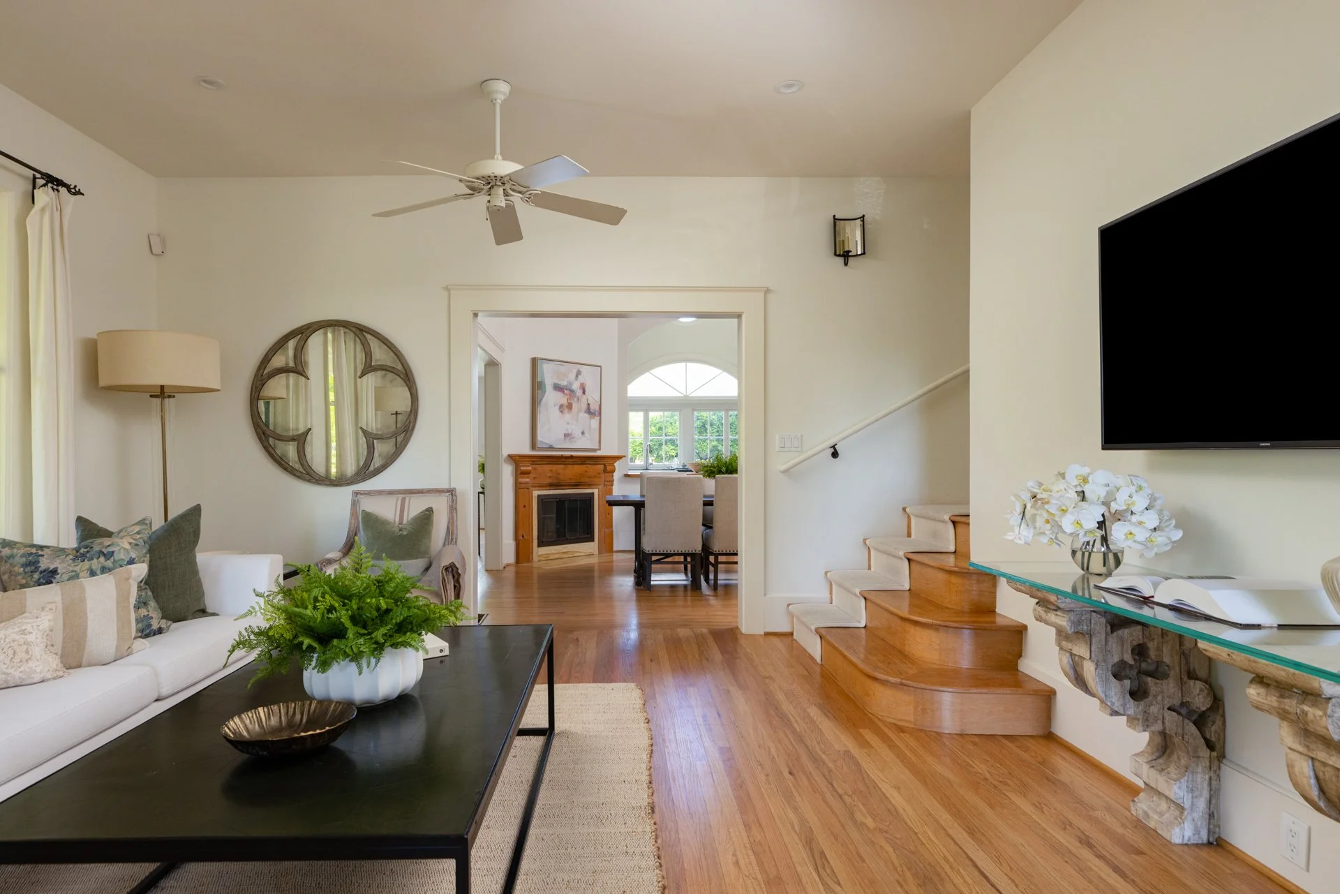 Living room with white sofa, green plants, wall mirror, wooden stairs, flat-screen TV, glass-top table with white flowers, and an adjoining dining and kitchen area in the background.