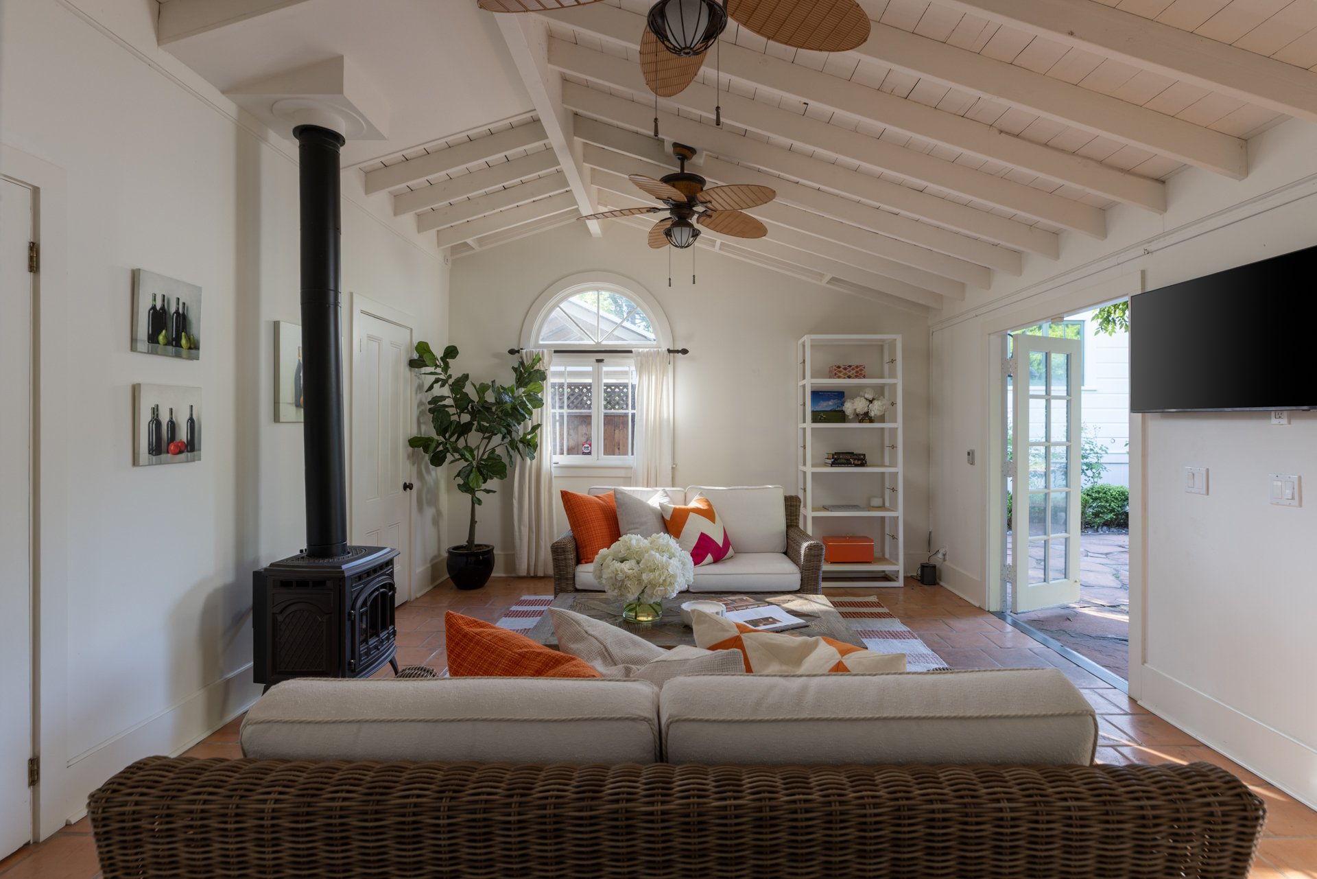 Living room with white walls, vaulted ceiling with white beams and ceiling fans, a black stove pipe, a white couch with orange and white pillows, a wicker armchair in front, a coffee table with a bouquet of white flowers, a potted plant, a window wit