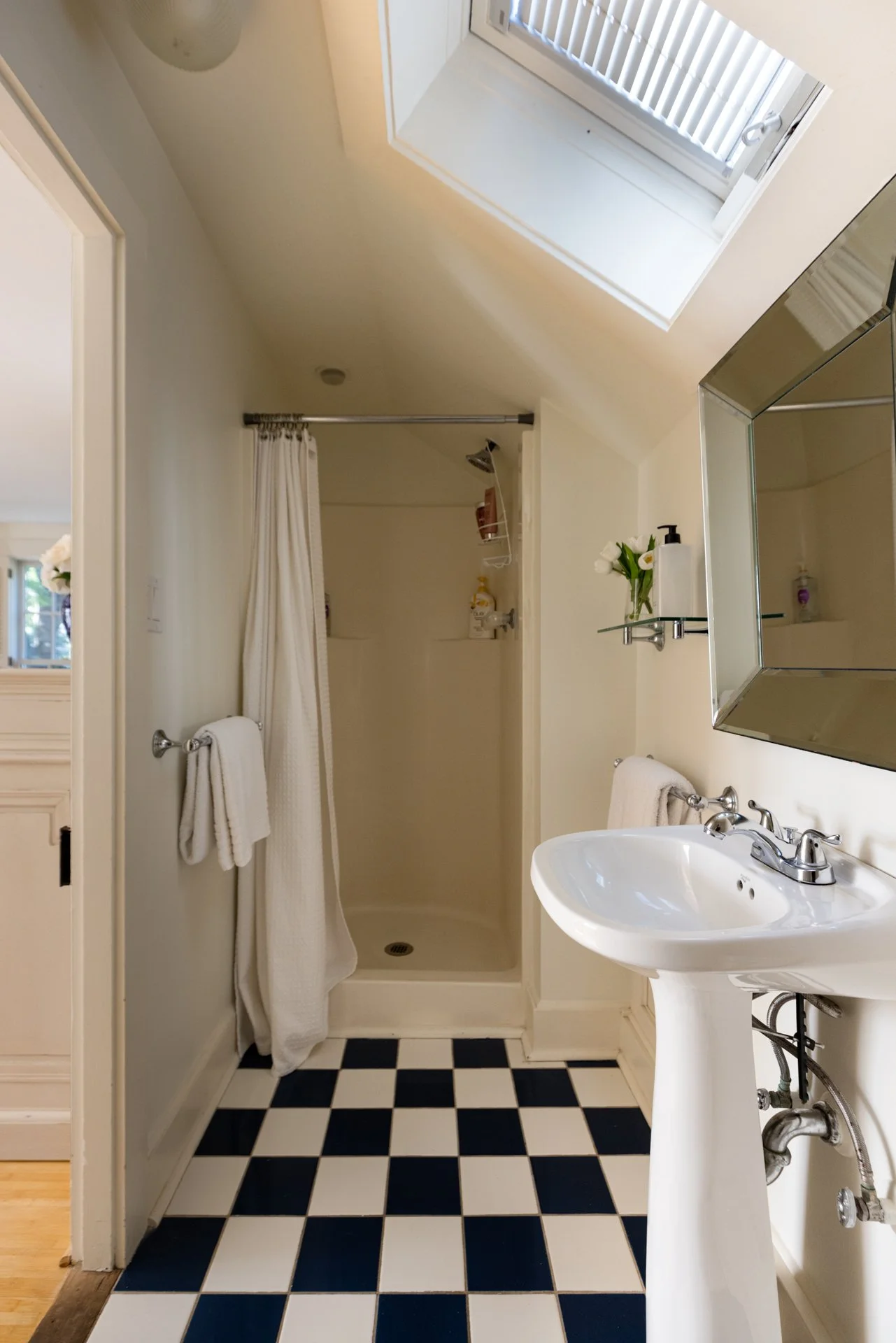 A bathroom with a black and white checkered floor, a white sink, a mirror cabinet, a small shelf with flowers, a shower area with a curtain, and a skylight window.