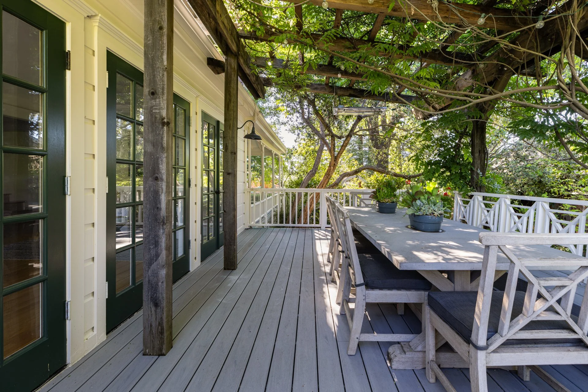 A spacious outdoor wooden deck connected to a house with yellow walls and green-framed glass doors, surrounded by trees and greenery. There is a large white wooden dining table with benches and a potted succulent plant, partly shaded by a trellis and