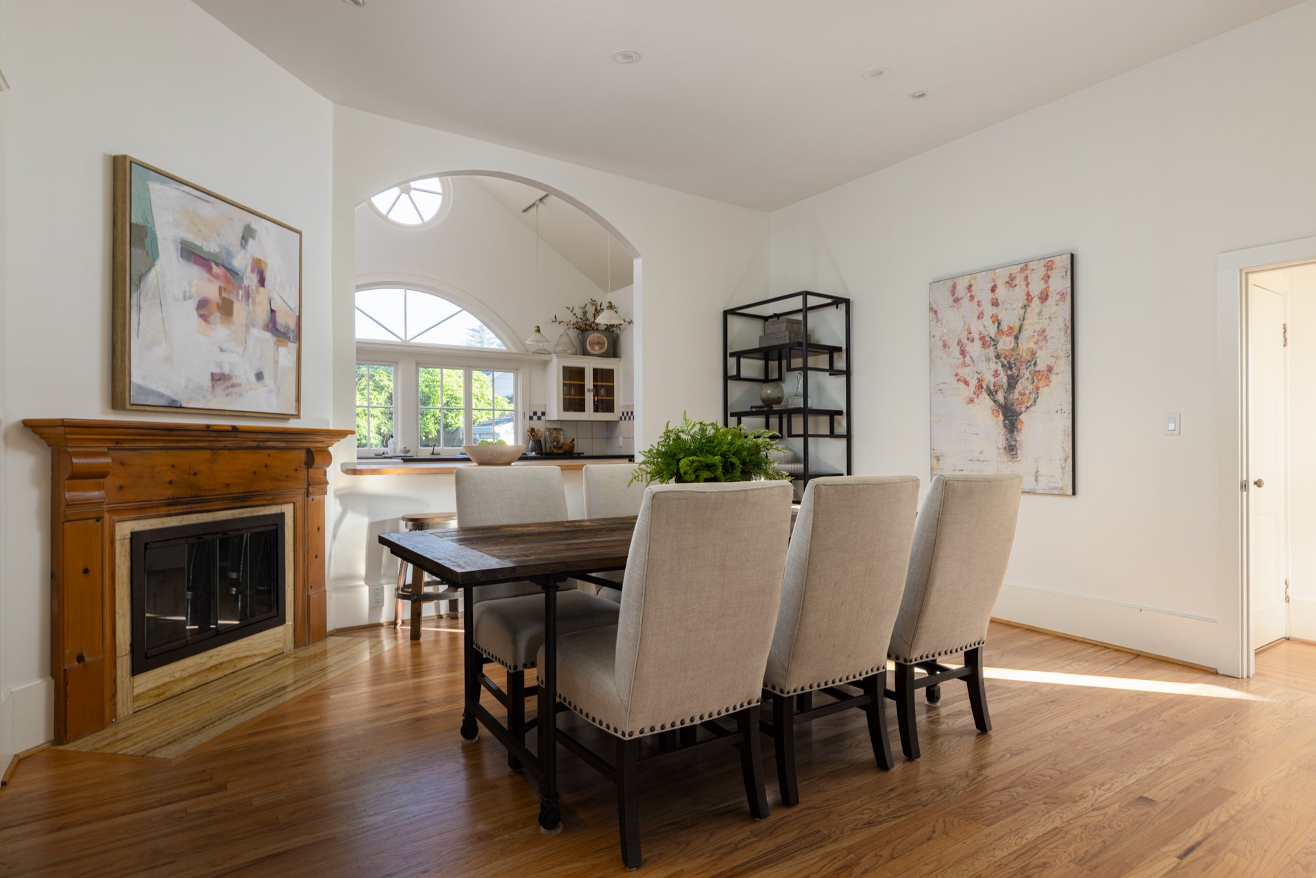 Interior view of a dining room with beige upholstered chairs around a wooden table, a black metal shelving unit, a fireplace with a wooden mantel, and artworks on white walls, with a kitchen visible through a large arched opening and windows letting 