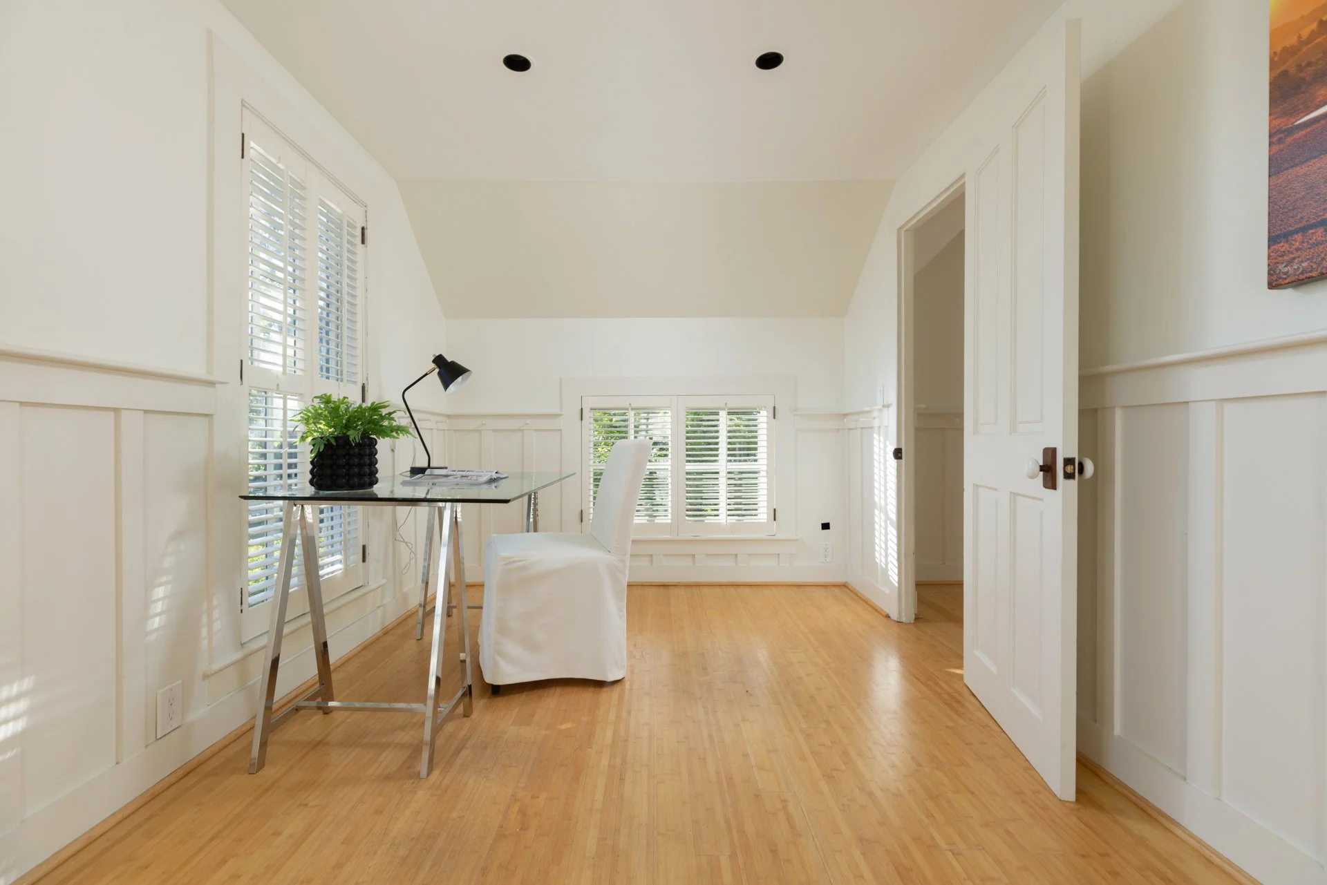 Bright room with hardwood floors, white walls with wainscoting, large windows with blinds, a glass desk with a plant and desk lamp, and a white chair.