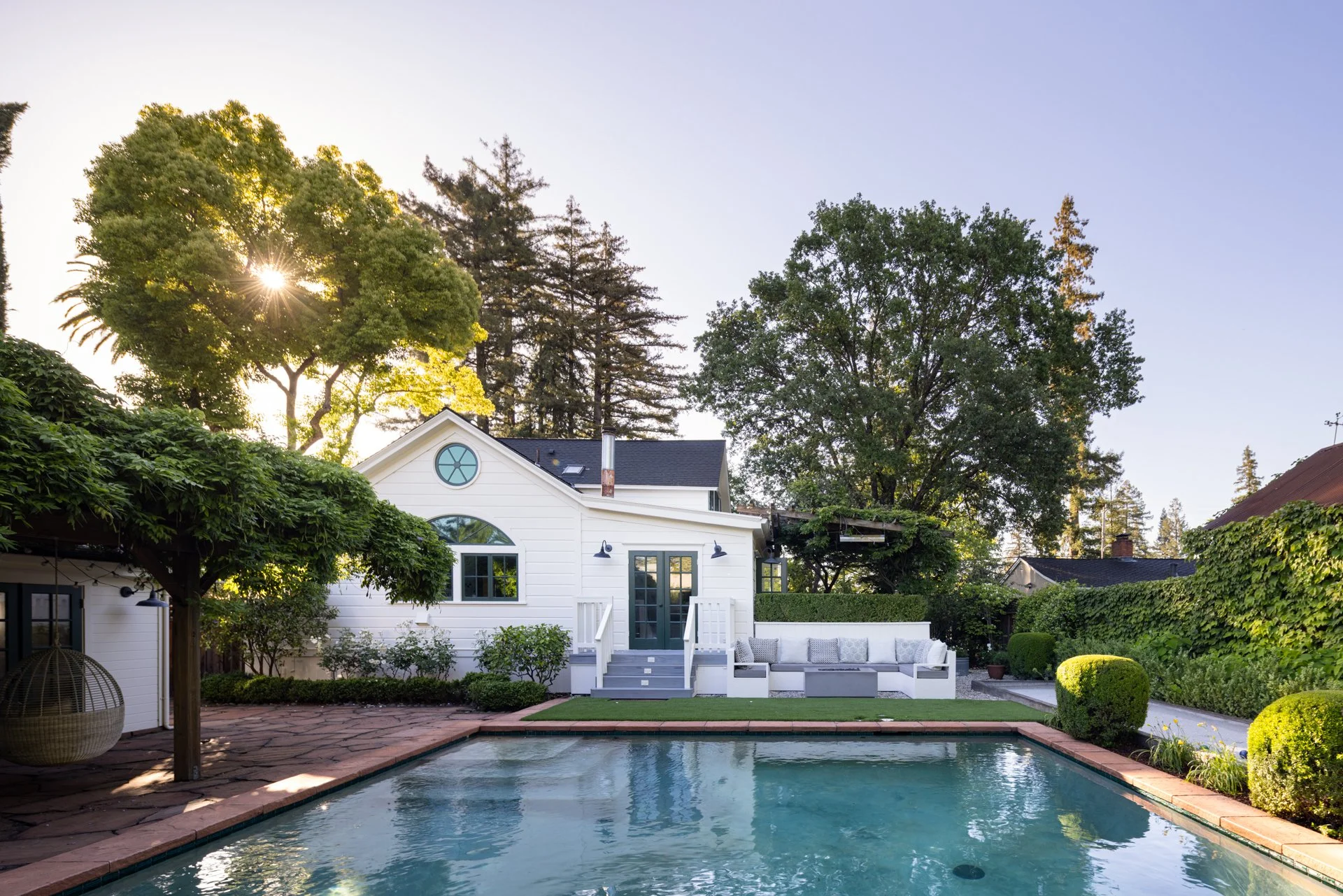 A backyard with a rectangular swimming pool, surrounded by greenery and a white house with dark doors and windows. Tall trees with sunlight shining through their leaves are in the background, and there is a patio area with outdoor seating.
