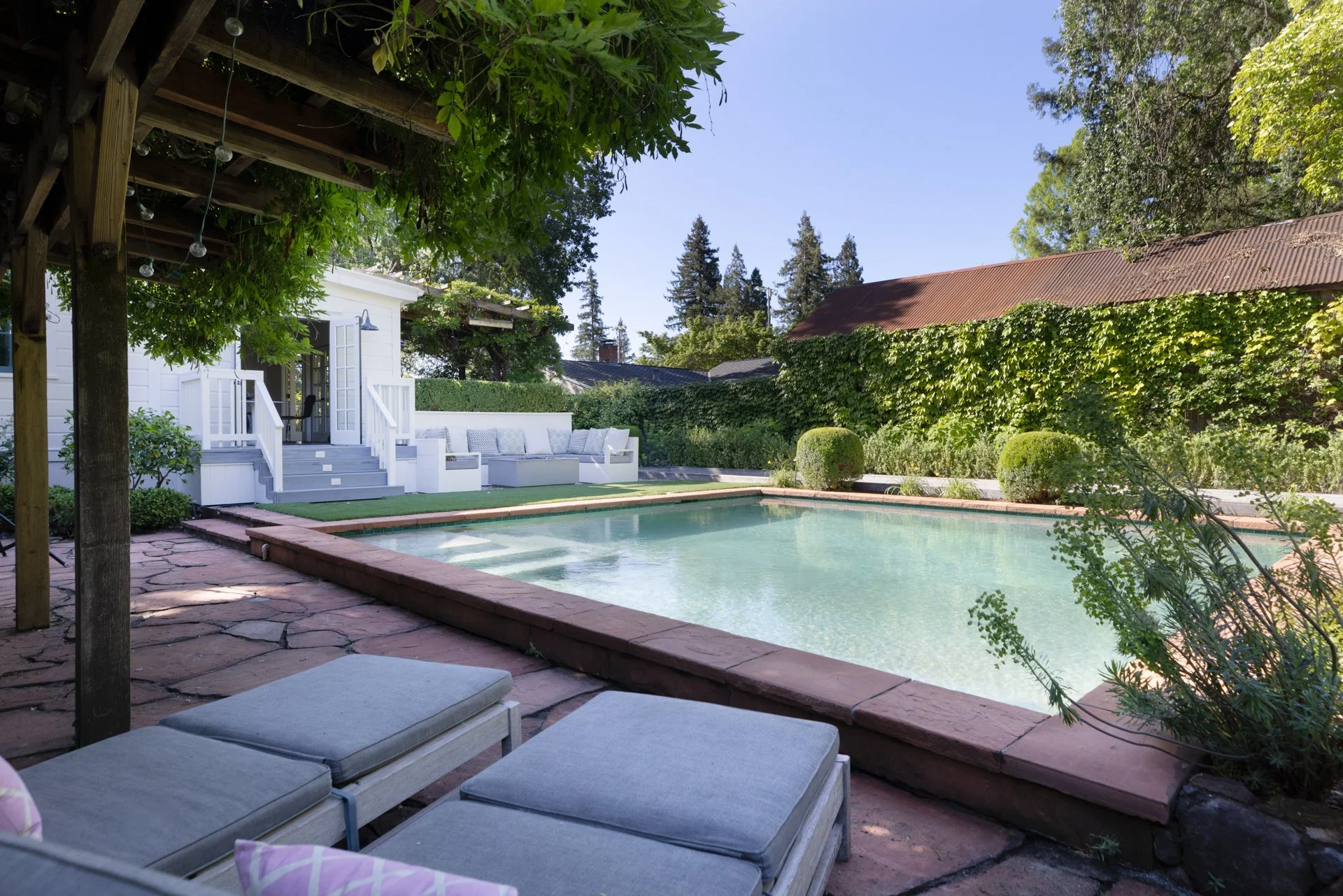 A backyard with a swimming pool surrounded by greenery, a white wooden deck with steps and cushions, and lounge chairs on a stone patio in the foreground. There are trees and a building with a metal roof in the background.