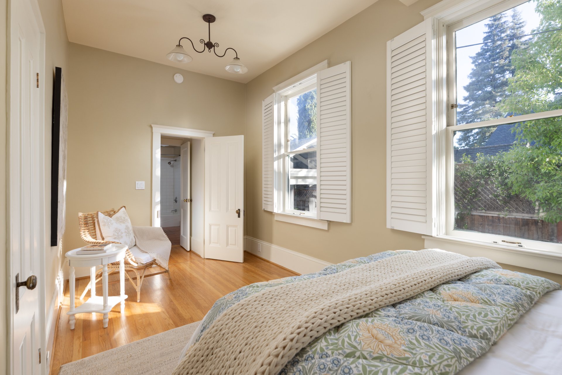 Bedroom with large windows, made bed with patterned sheets, corner with white chair and side table, open white door leading to bathroom, hardwood floors, neutral walls, ceiling light fixture.