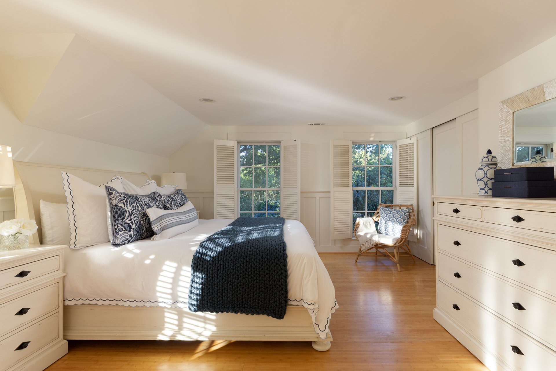 Bright bedroom with a white bed, navy blanket, and decorative pillows, three windows with white shutters, a wicker chair, and a cream dresser with a mirror and decorative items.