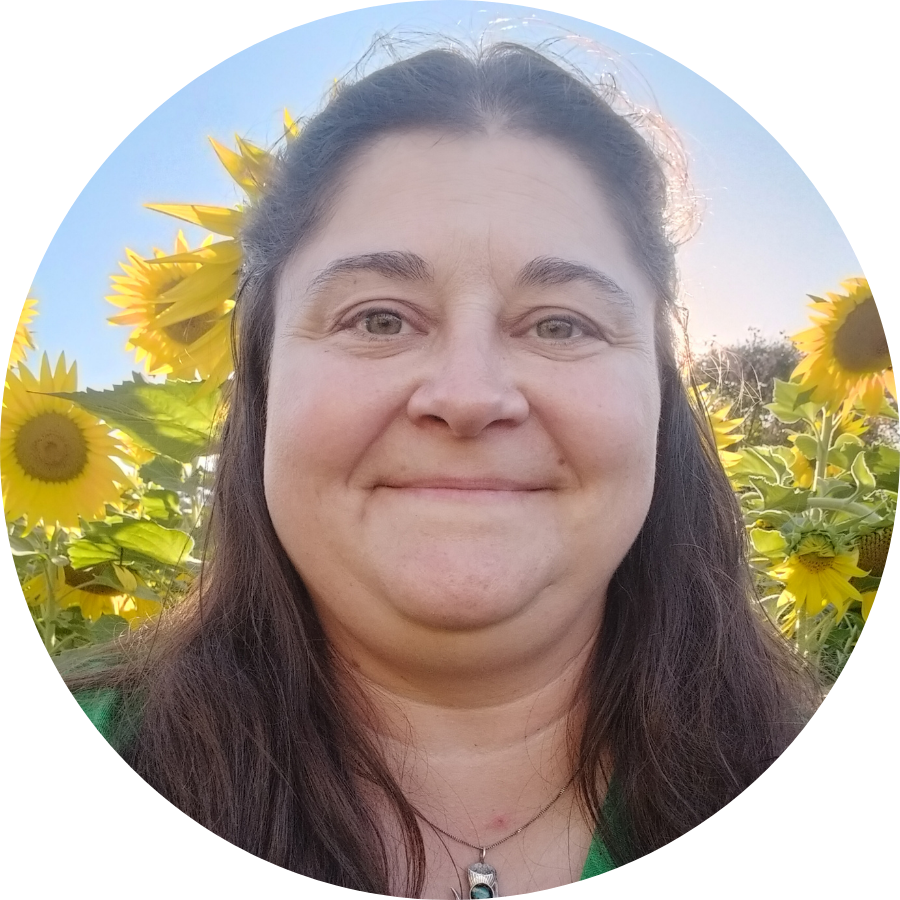 A woman with brown hair smiling outdoors in a sunflower field with blue skies in the background.