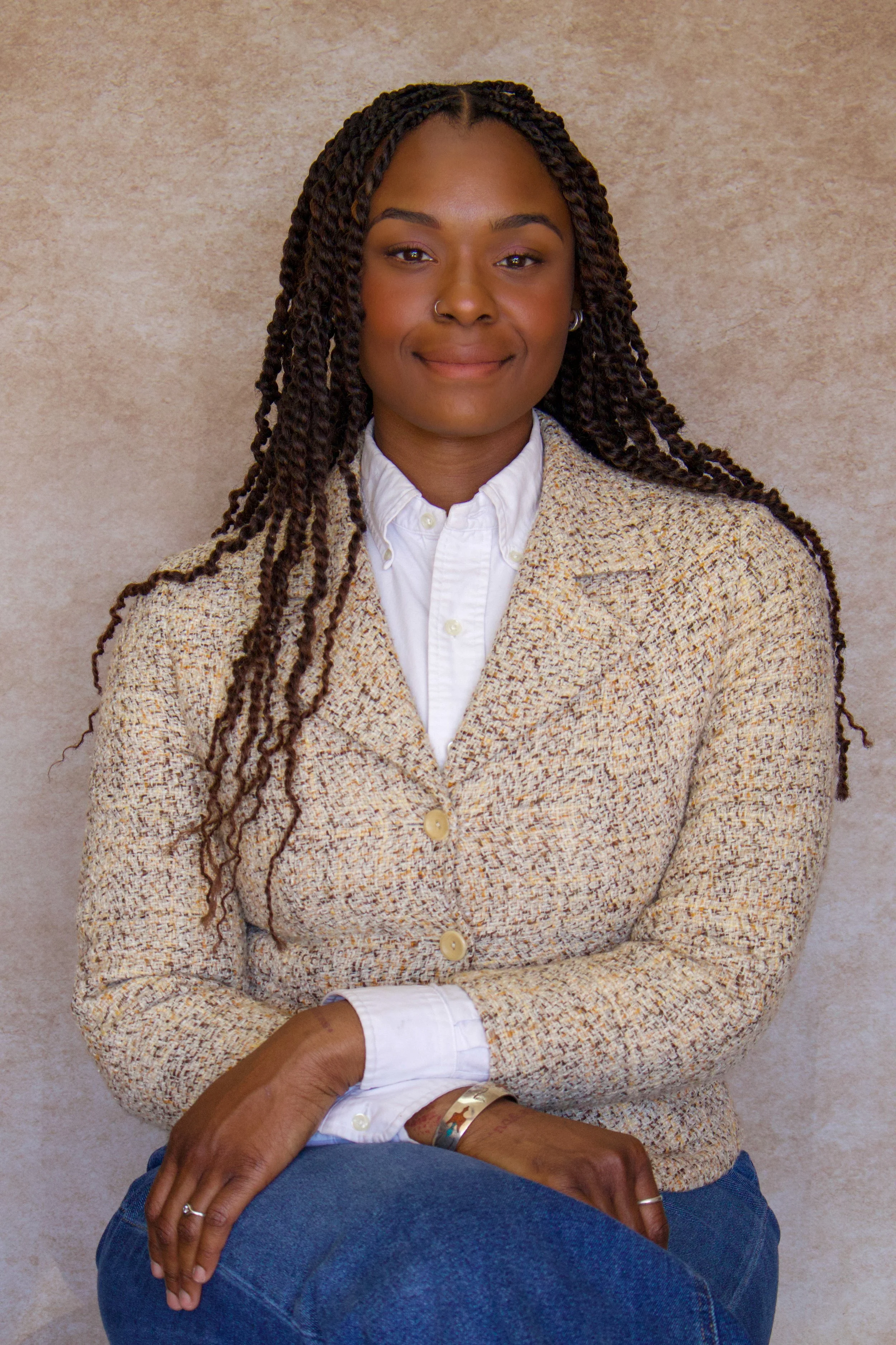 Portrait of a young woman with brown skin, braided hair, and a nose ring, wearing a white shirt and a beige tweed jacket, sitting with her arms crossed, smiling gently against a beige textured background.