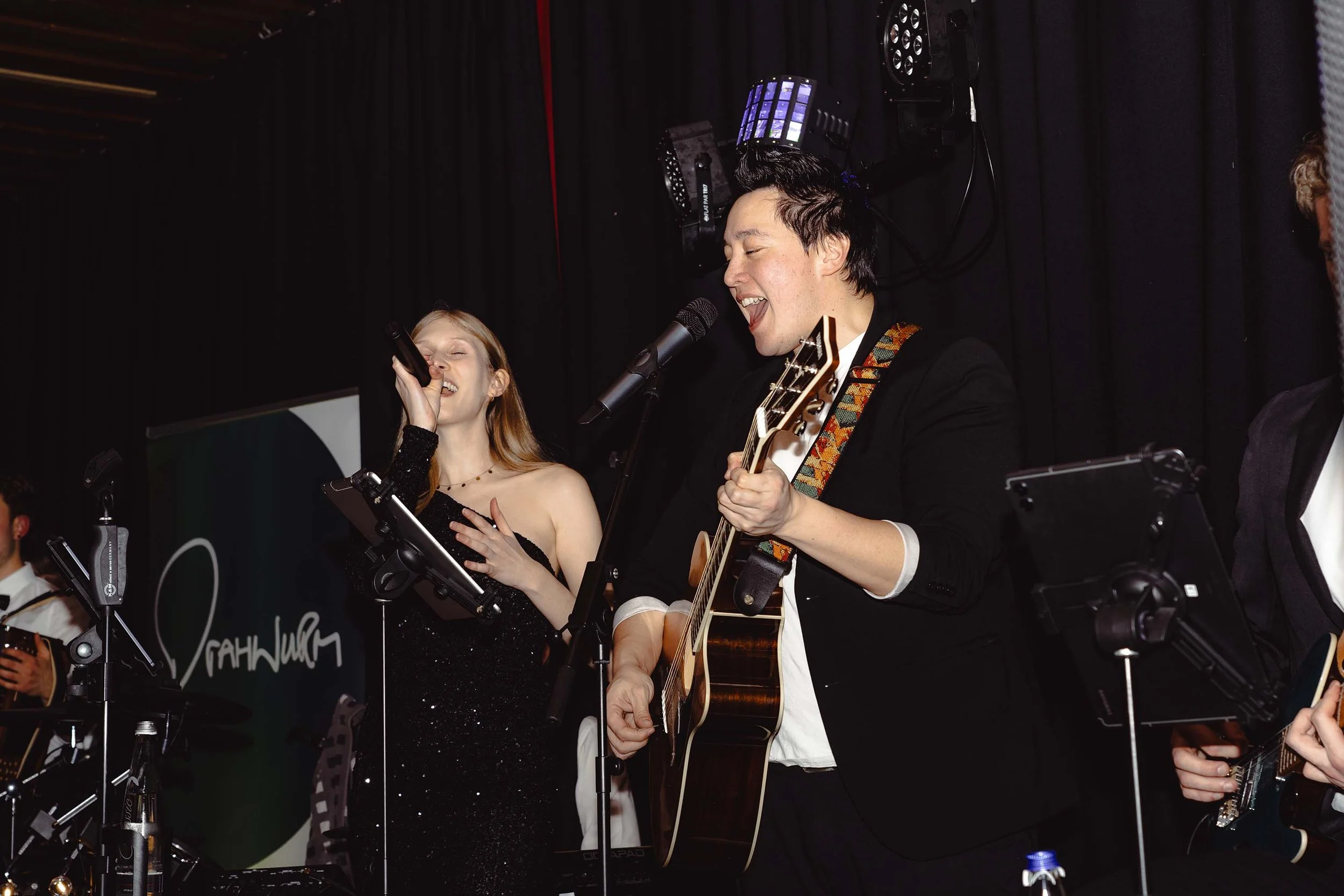 Musiker beim Auftritt, singend und Gitarre spielend, in einem dunklen Raum mit schwarzen Vorhängen im Hintergrund.