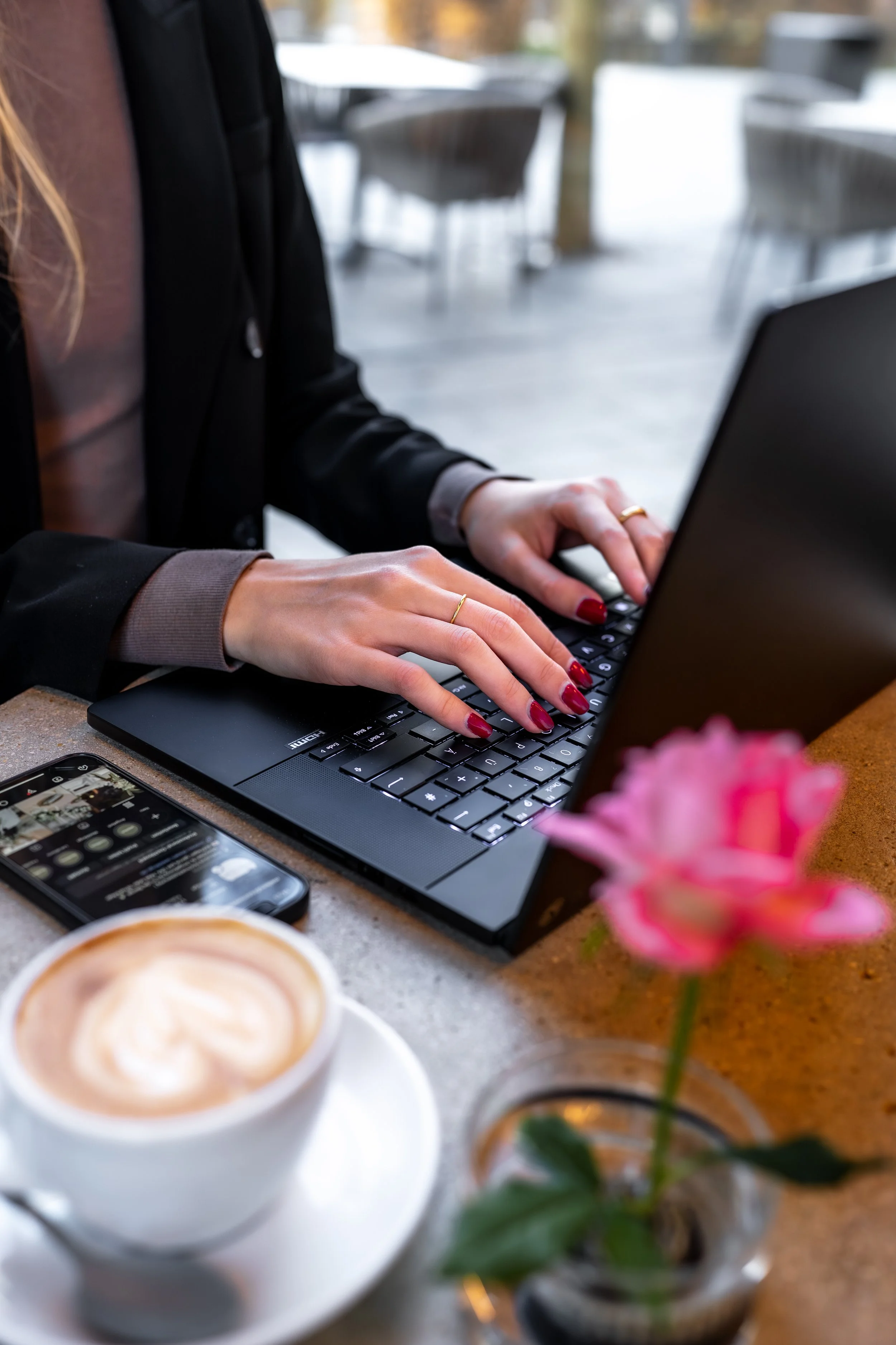 Nahaufnahme einer Person, die auf einem Laptop tippt, mit einem Kaffee und einem Smartphone auf einem Tisch, im Hintergrund sind leere Stühle zu sehen, im Vordergrund eine pinke Blume.