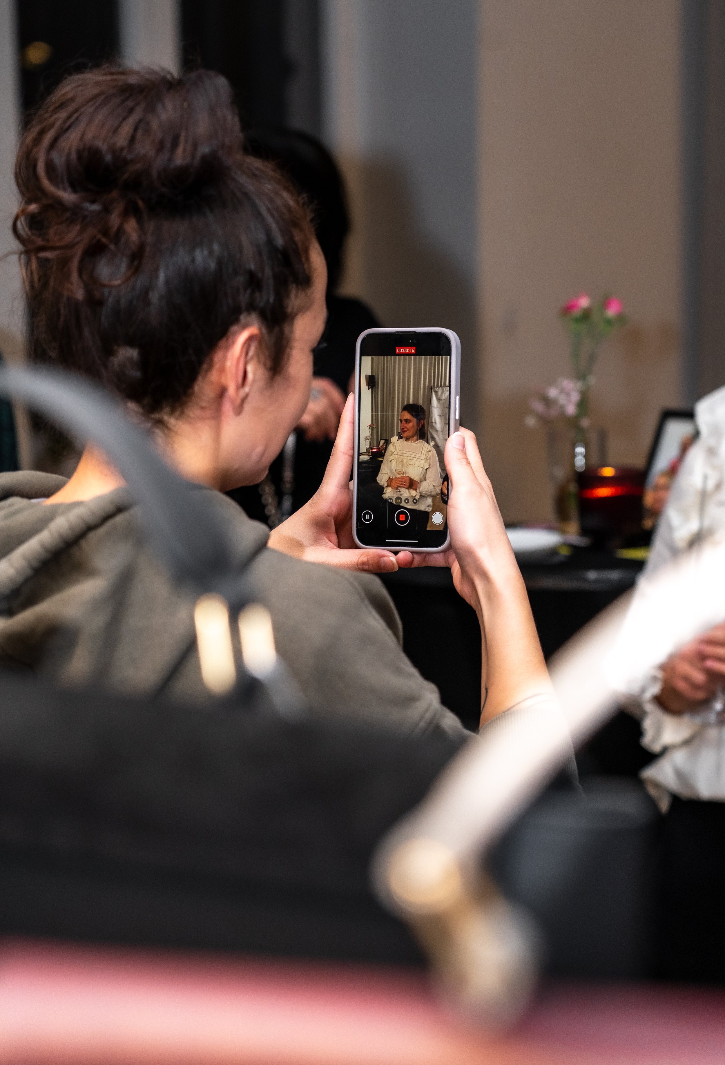 Person filmt eine Frau in einem weißen Kleid mit einem Smartphone bei einer Veranstaltung.