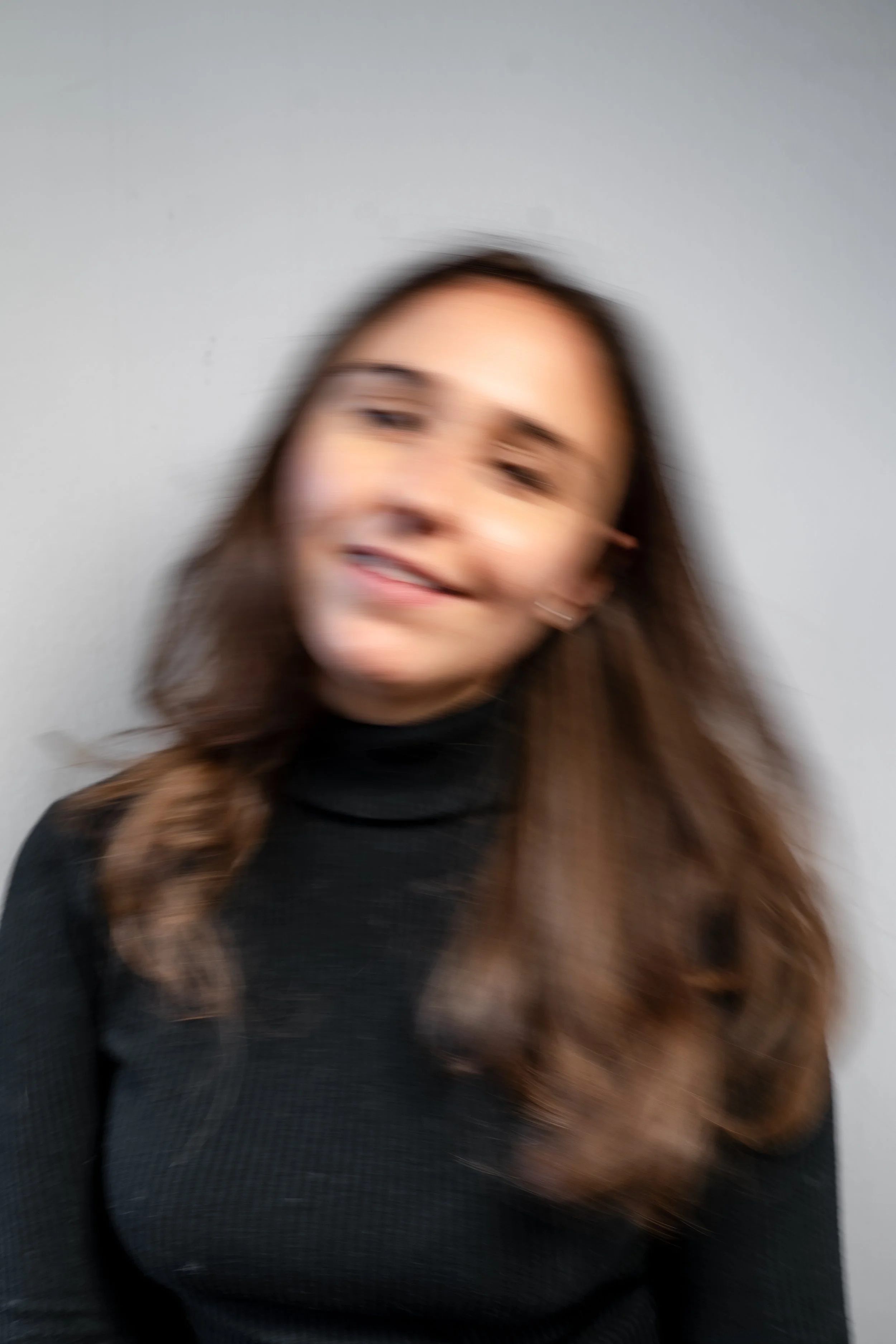 Das Bild zeigt eine junge Frau mit brünettem, länglichem Haar, die vor einem neutralen grauen Hintergrund steht. Sie trägt einen schwarzen, langärmligen Pullover.